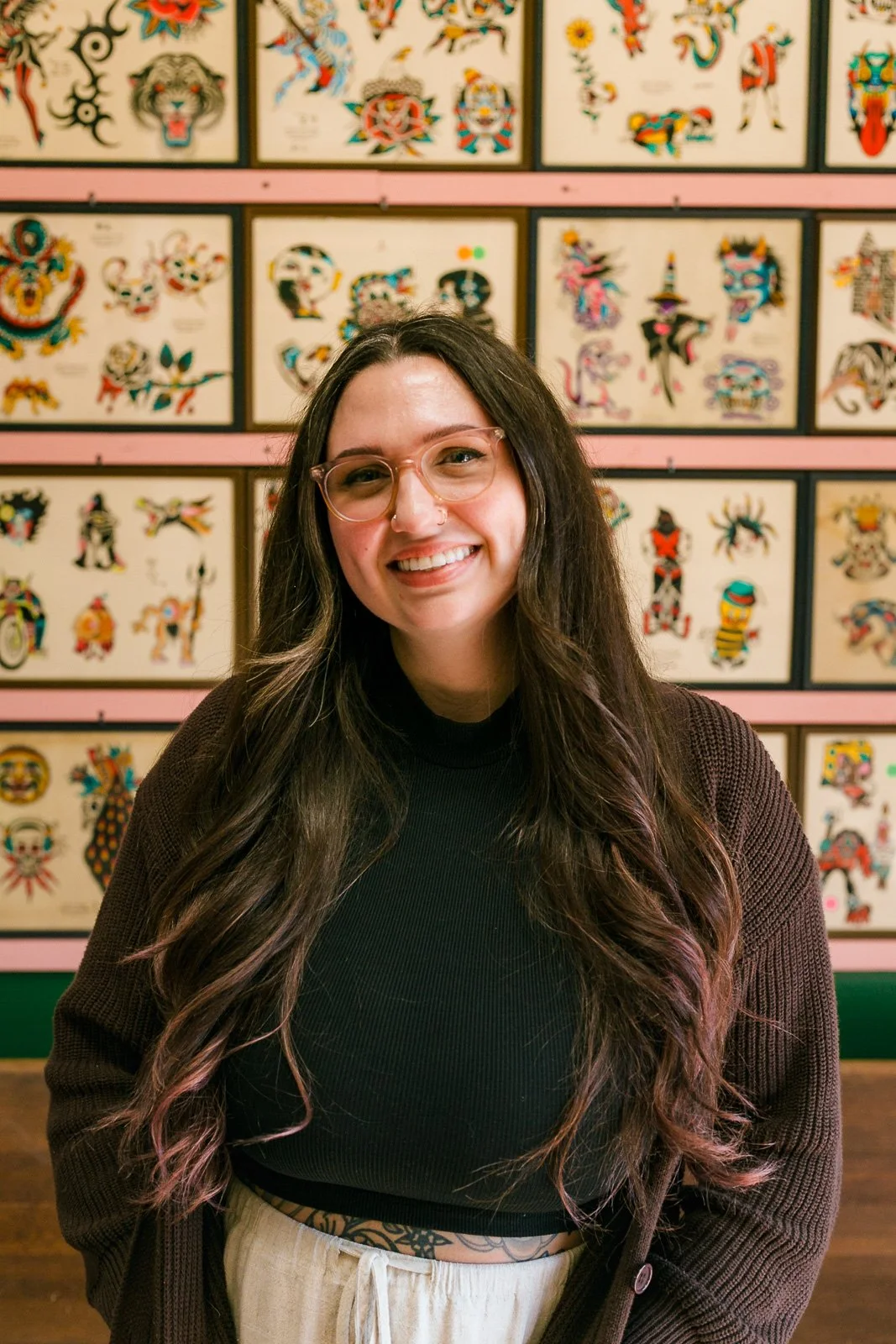 A woman with long dark hair, glasses, and a nose ring smiling in front of a colorful tattoo flash wall.