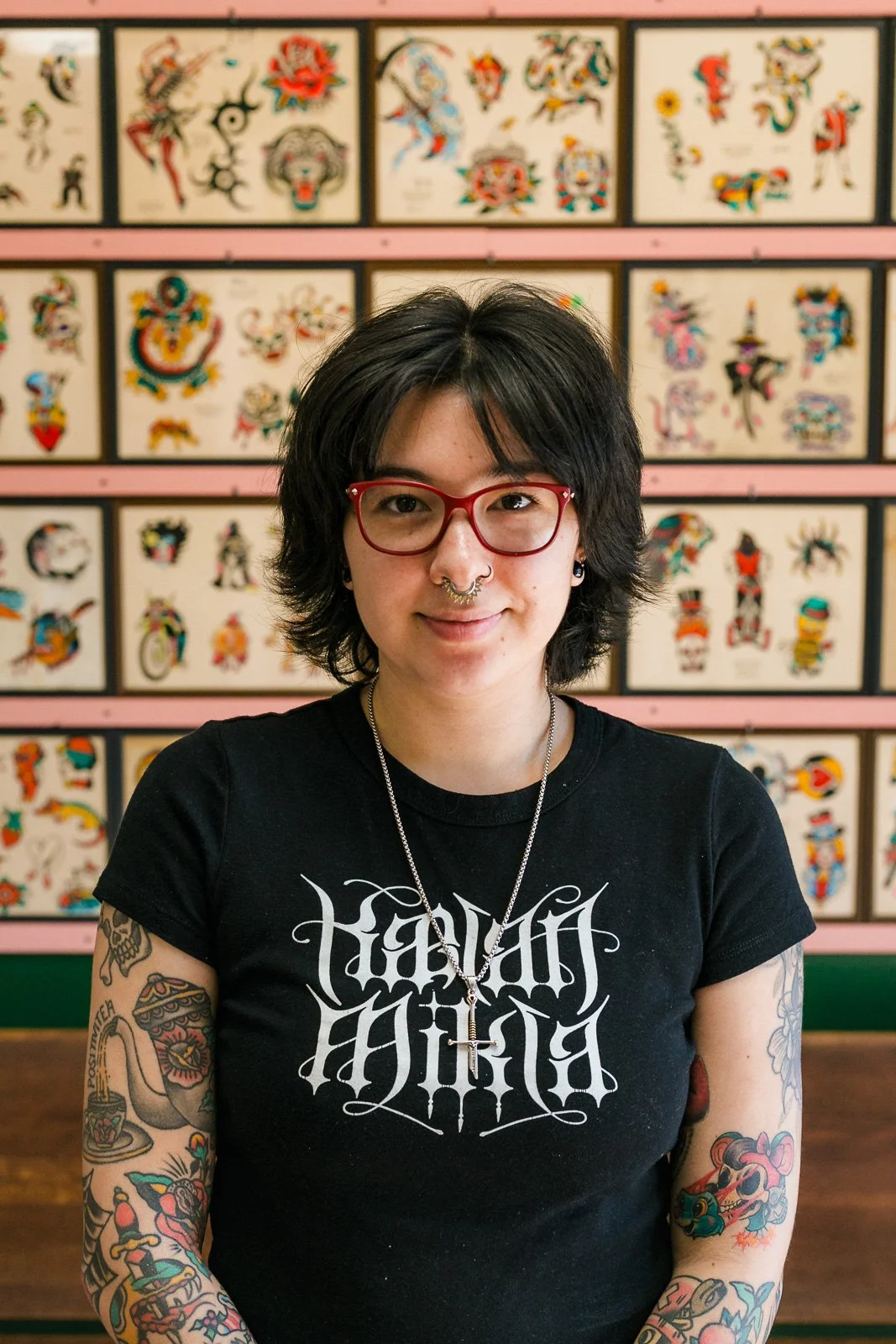 A woman with black hair, red glasses, and tattoos on her arms, wearing a black T-shirt with white gothic text, sitting in front of a wall decorated with colorful tattoo flash art.