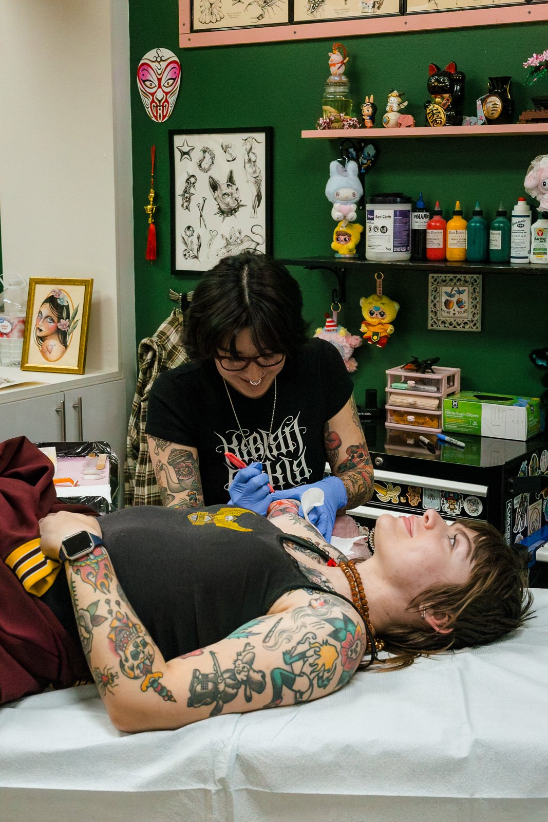 A woman with tattoos lying on a tattoo chair, getting a tattoo from a tattoo artist. The artist has black hair, glasses, and tattoos on her arms, and is wearing blue gloves. The room has colorful decorations, artwork, and tattoo supplies on shelves.