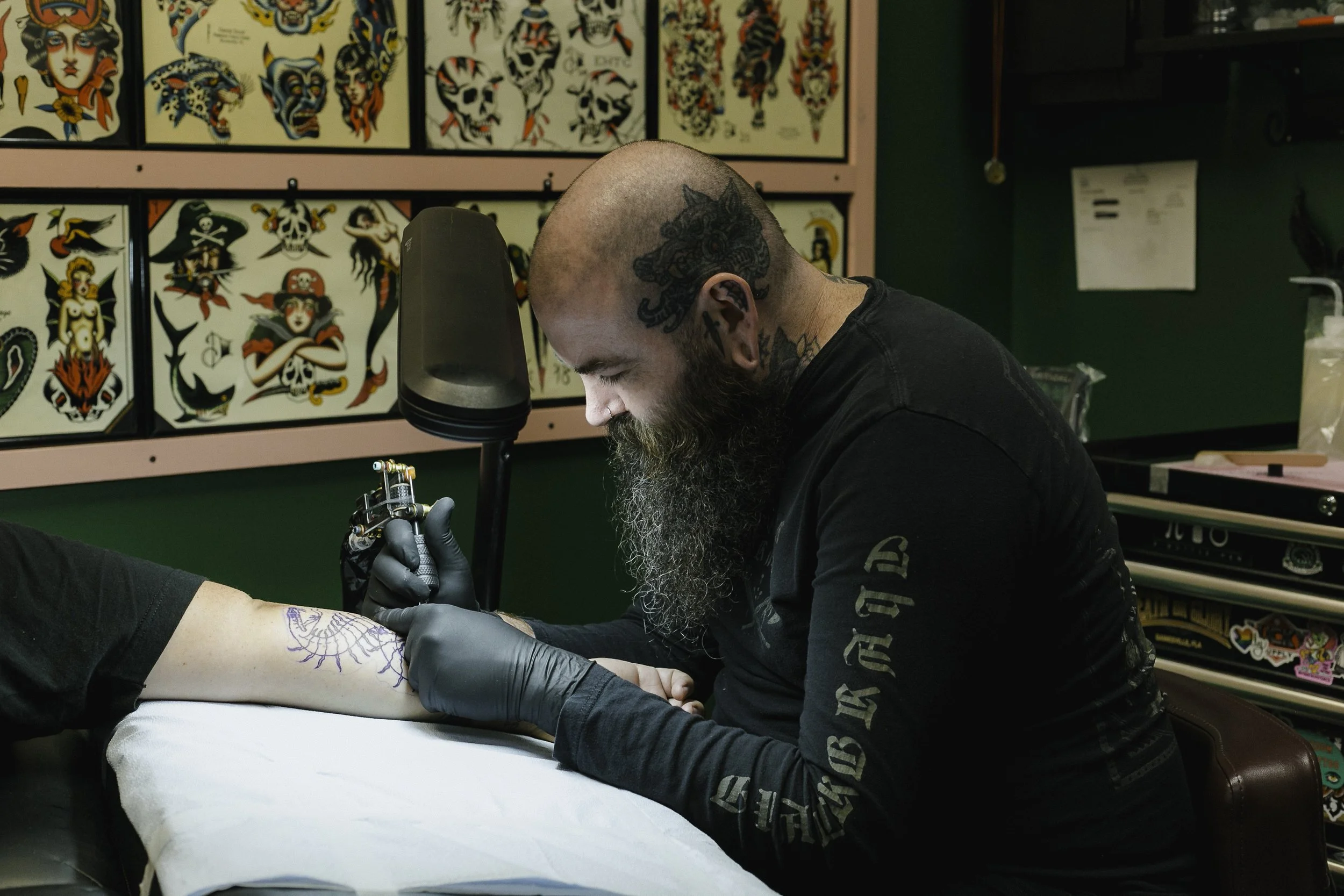 A tattoo artist working on a tattoo on a person’s forearm in a tattoo shop decorated with traditional tattoo flash art. The tattoo artist has a beard, tattoos on his head and neck, and is wearing gloves while focused on his work.