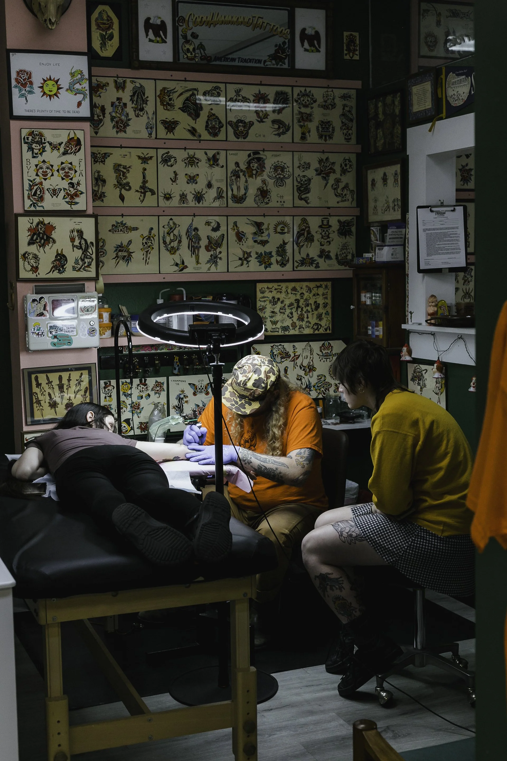 A tattoo studio with walls decorated with various tattoo flash designs. Two clients are lying on a tattoo table, receiving tattoos while two artists work on them. The studio has a pink and green color scheme, with framed tattoo designs on the wall an