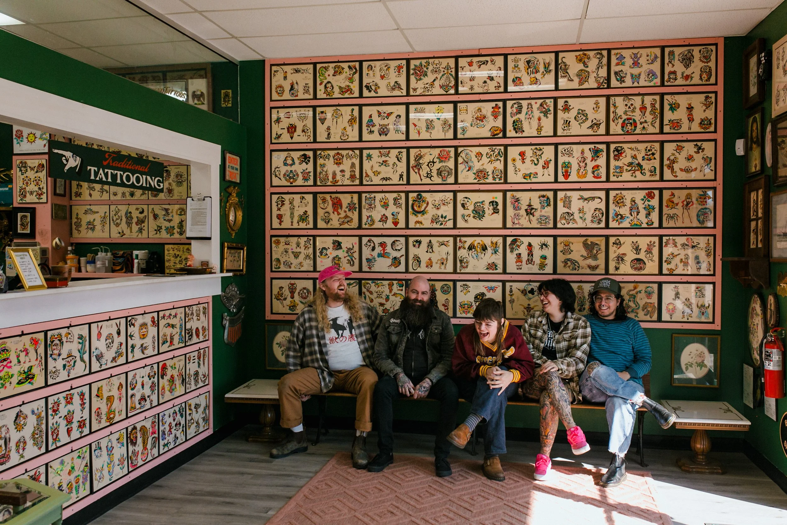 Group of five people sitting in front of a wall filled with colorful tattoo flash art at a tattoo shop, smiling and laughing.