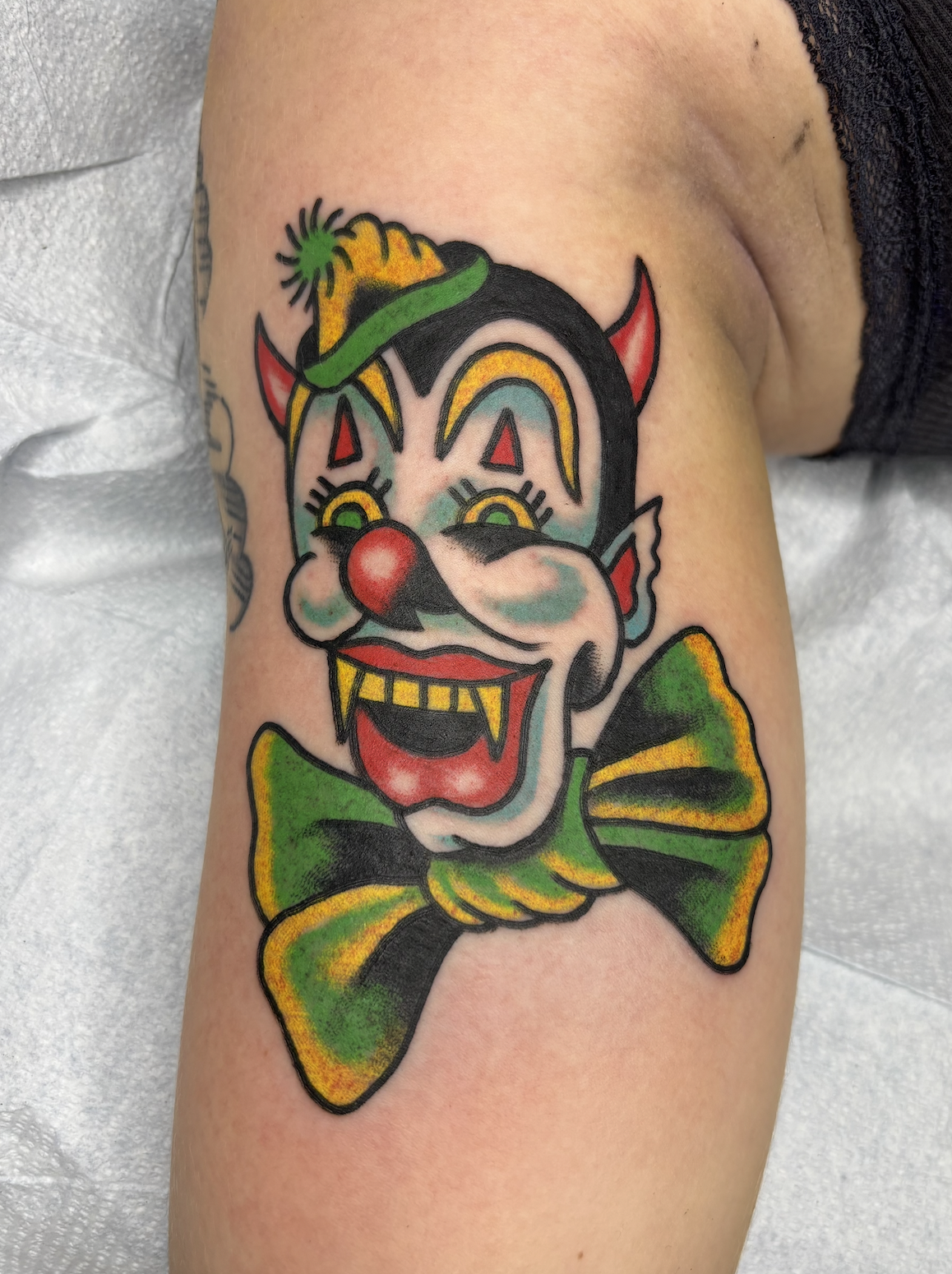 Colorful tattoo of a clown with a painted face, red nose, and yellow teeth, wearing a green bow tie and a hat with a yellow pom-pom, on person's upper arm.