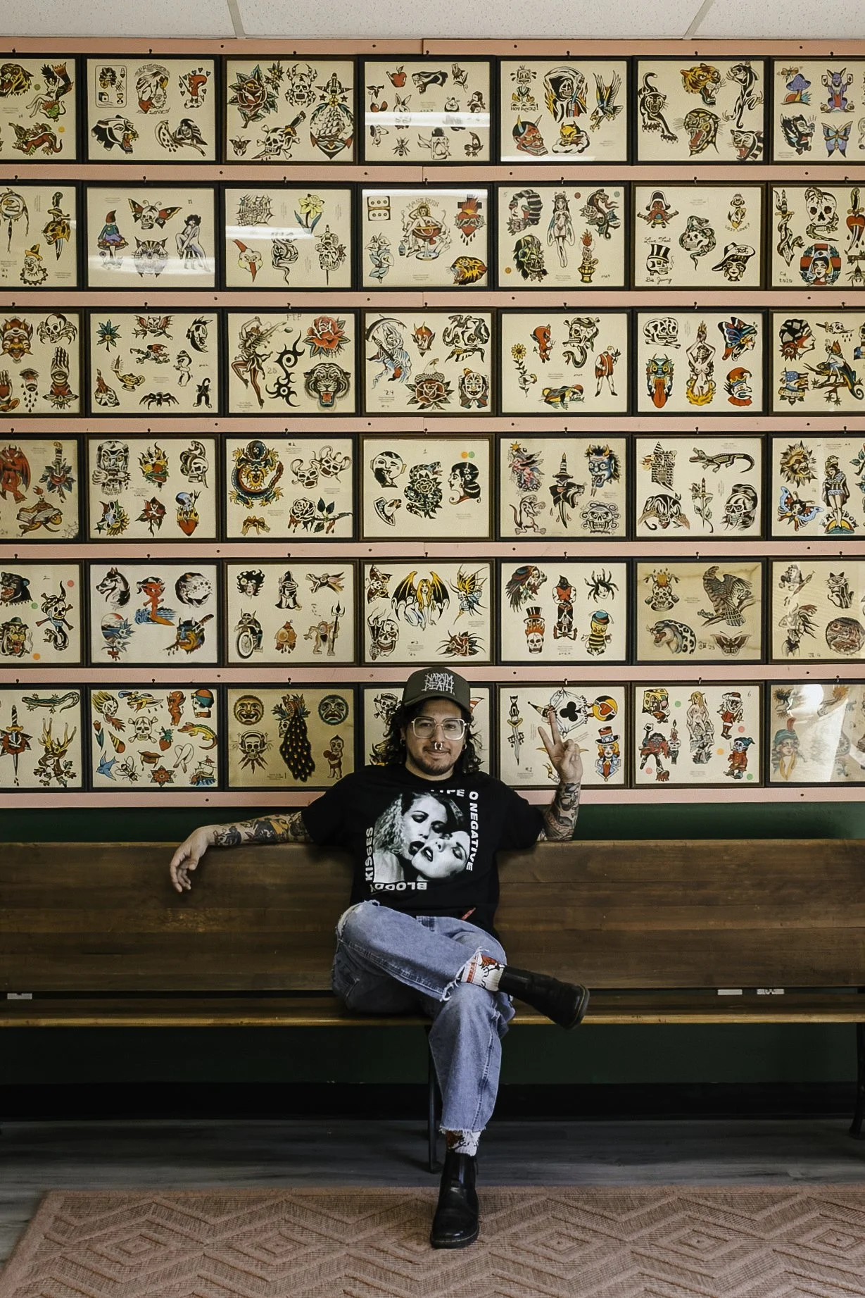 A man with tattoos sitting on a wooden bench in front of a wall of colorful tattoo flash art, making a peace sign with his right hand, wearing glasses, a black graphic t-shirt, ripped jeans, and black boots.