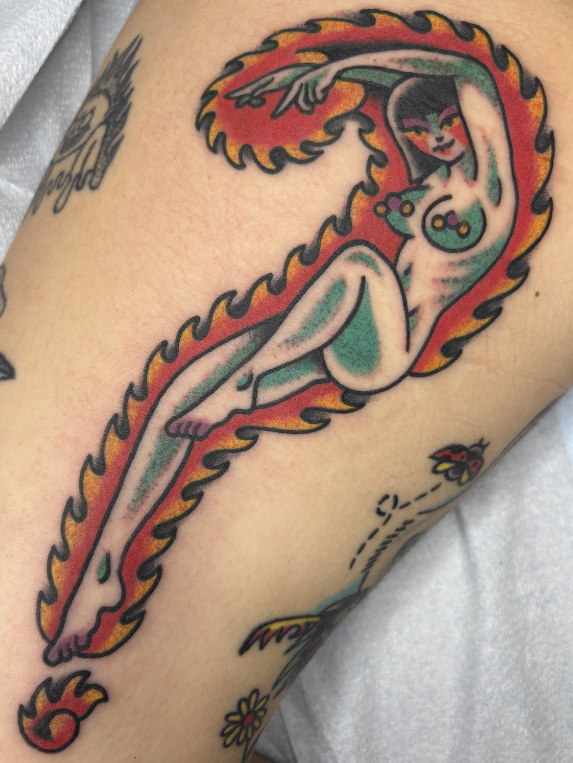 A colorful tattoo of a woman with dark hair and makeup, lying within a flame-like fiery border with green and red accents, on a person's upper arm.
