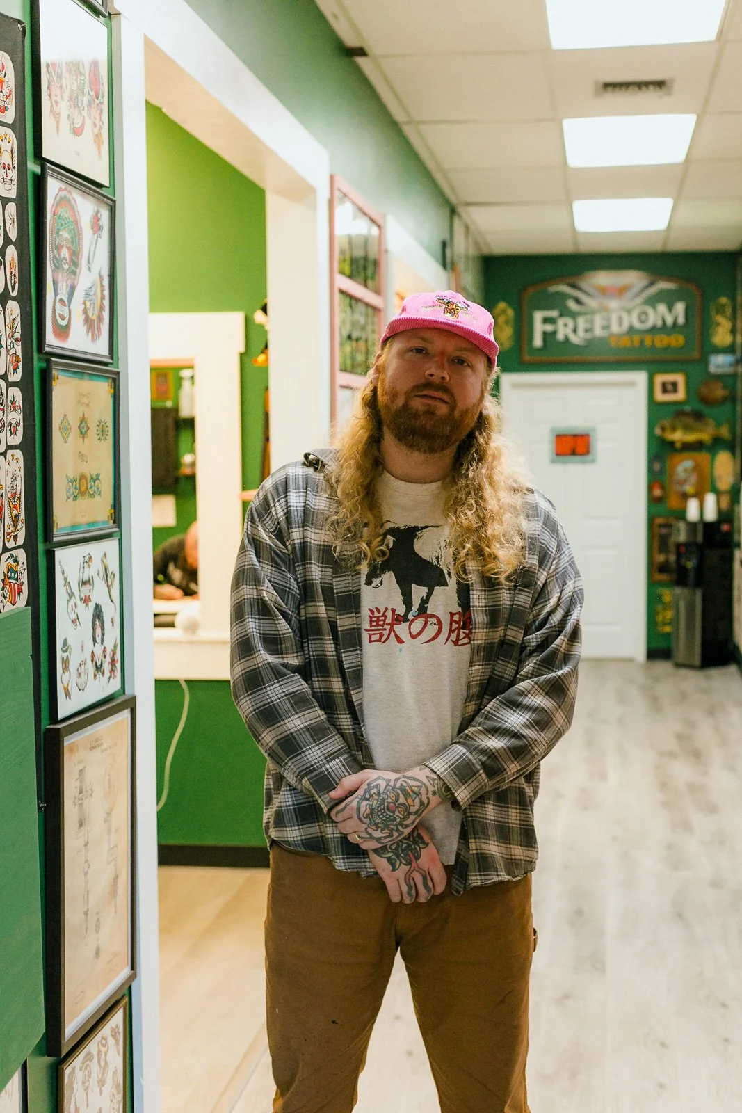 A man with long curly hair and tattoos on his hands poses in an indoor tattoo shop. He wears a pink baseball cap, a gray T-shirt with a black dog print and Japanese characters, a plaid button-up shirt, and brown pants. The shop has green walls, framed tattoo artwork on display, and a counter in the background.