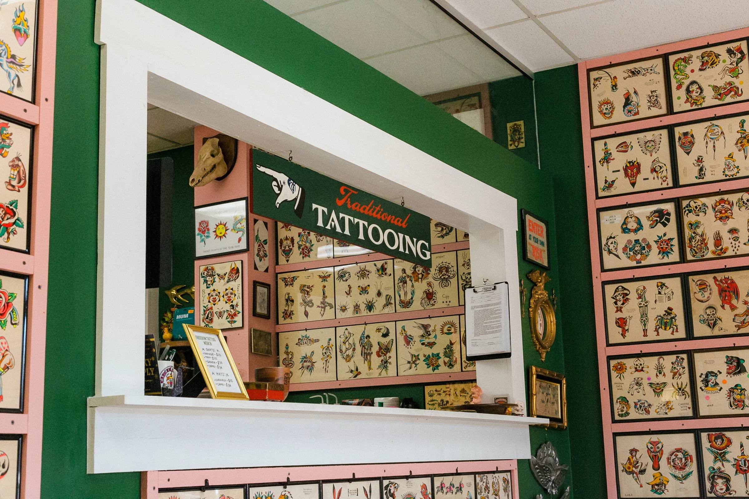 Interior of a tattoo shop with walls covered in colorful tattoo designs and flash art, a sign for traditional tattooing, and a butterfly and bird decorative clock.
