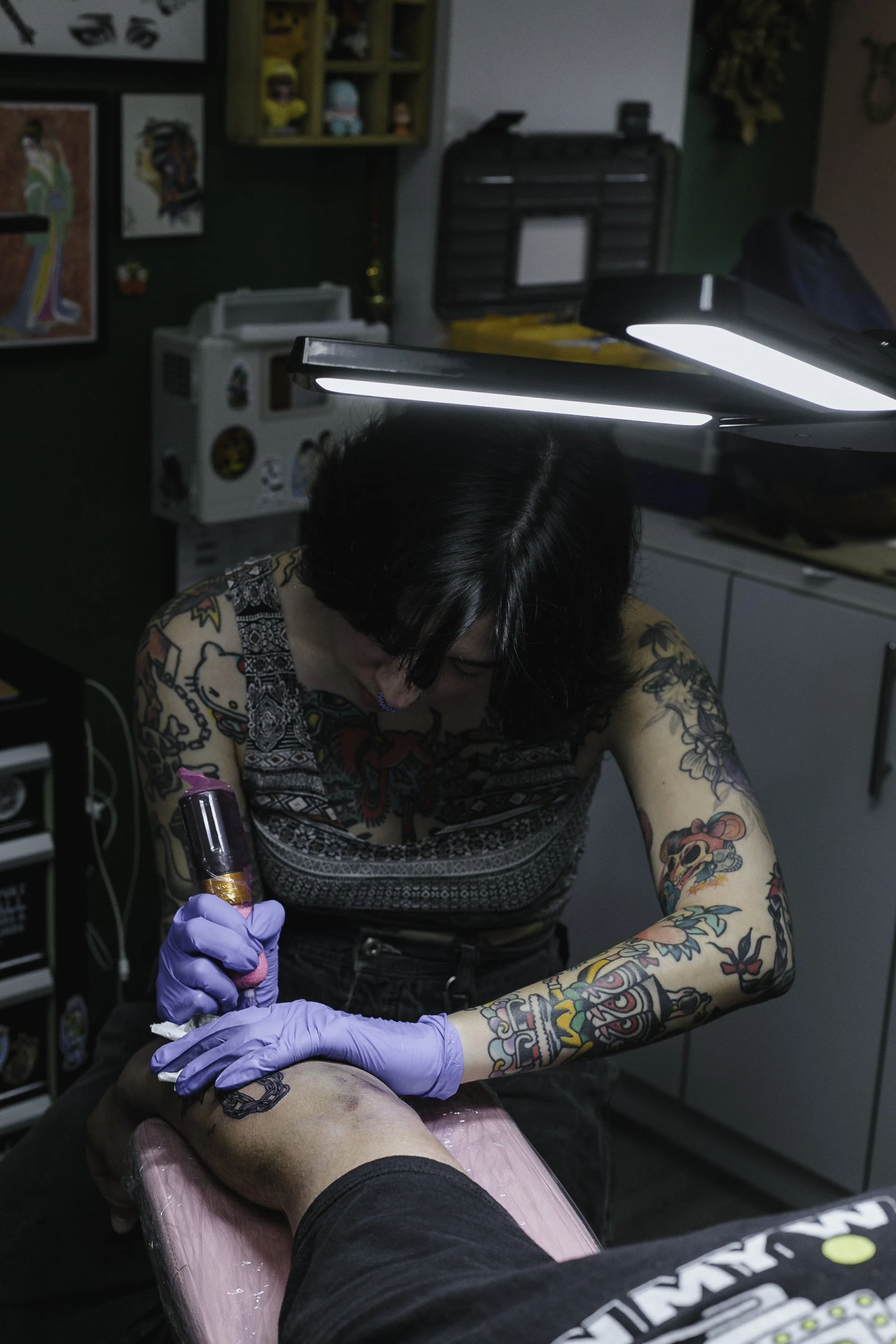 A tattooed woman performing a tattoo on a person's leg at a tattoo parlor, with tattoo equipment and artwork visible in the background.