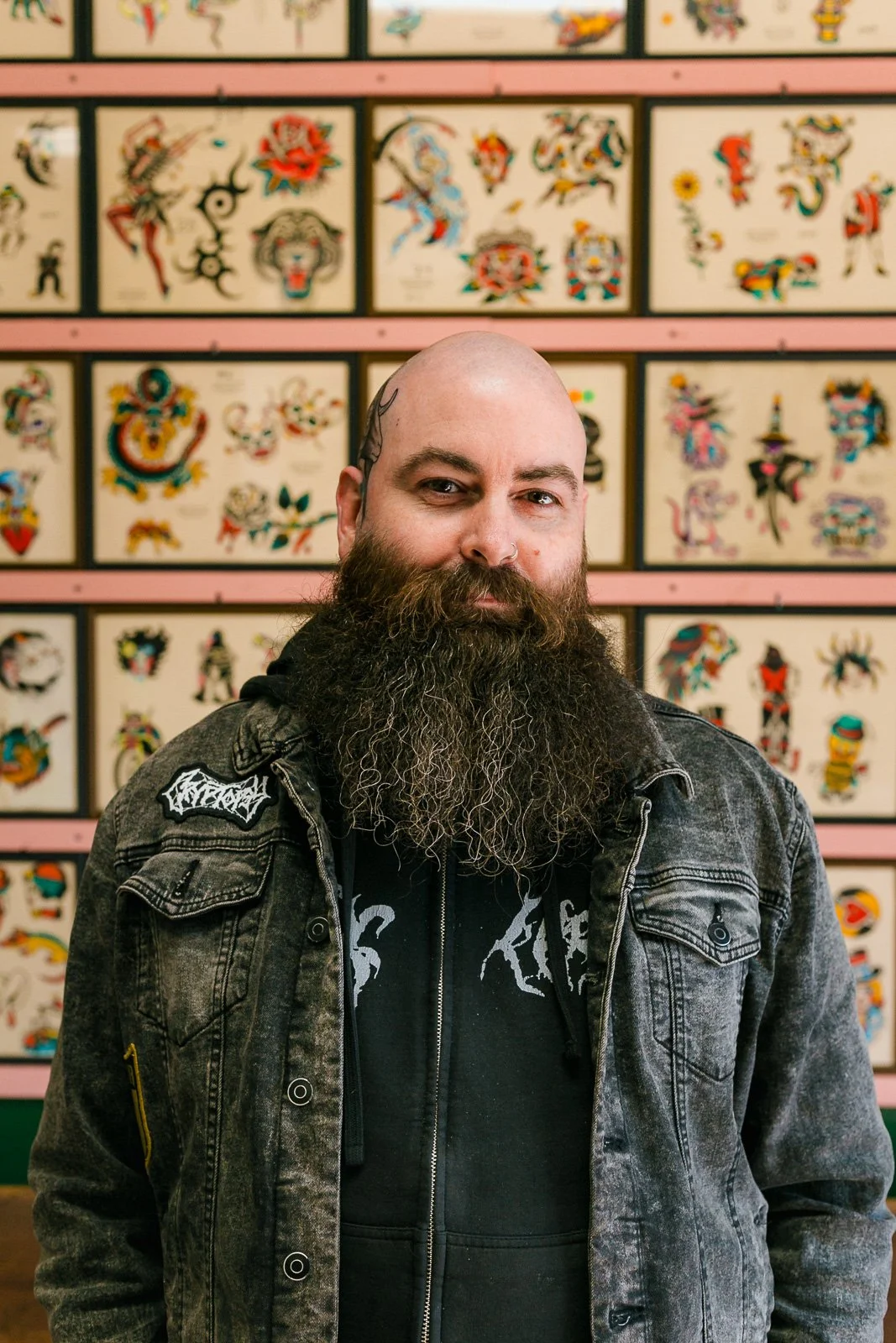 A man with a beard and tattoos on his head standing in front of a wall decorated with framed tattoo designs.