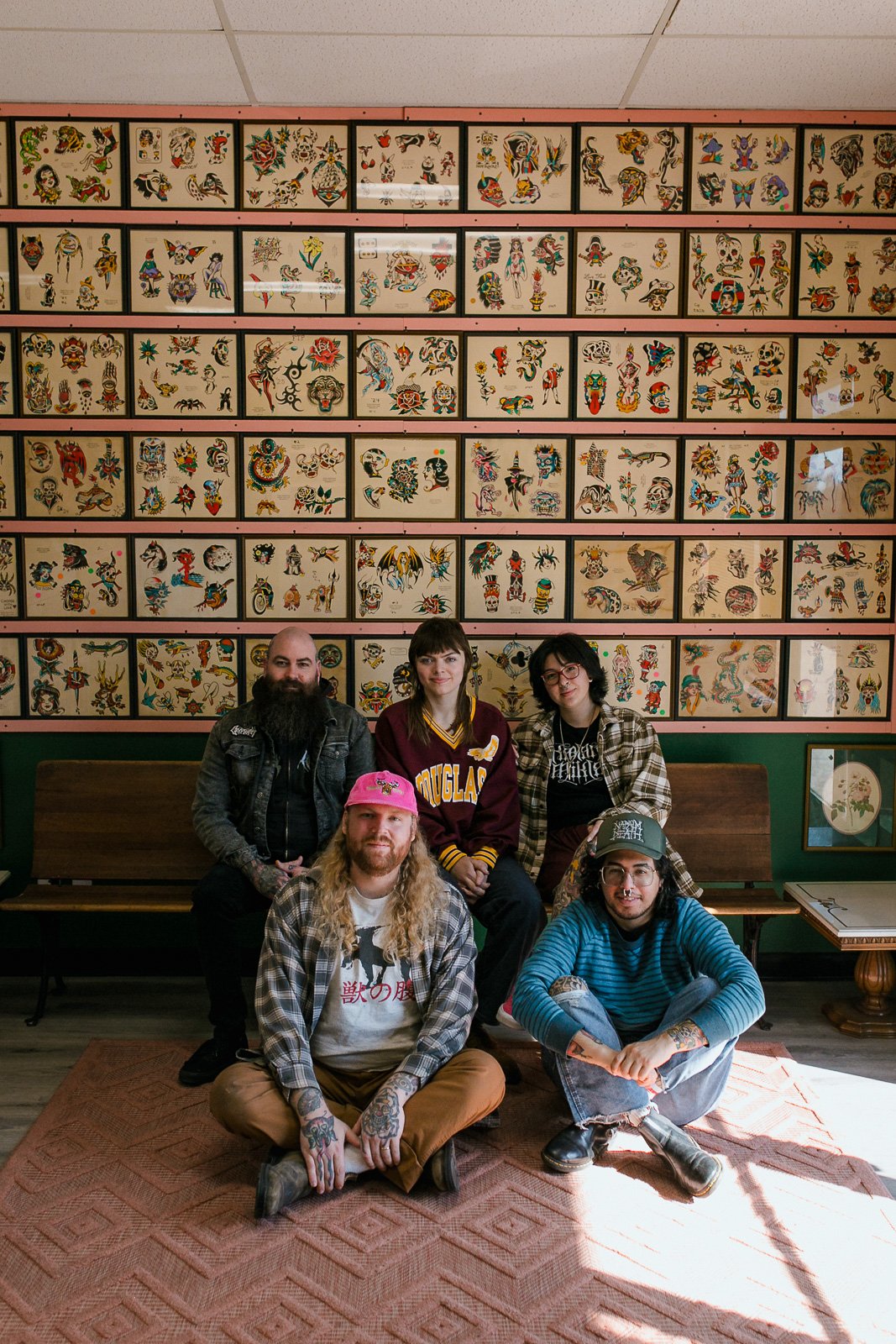 A group of five young adults sitting and standing in front of a wall decorated with framed tattoo flash art, in a tattoo shop or gallery.
