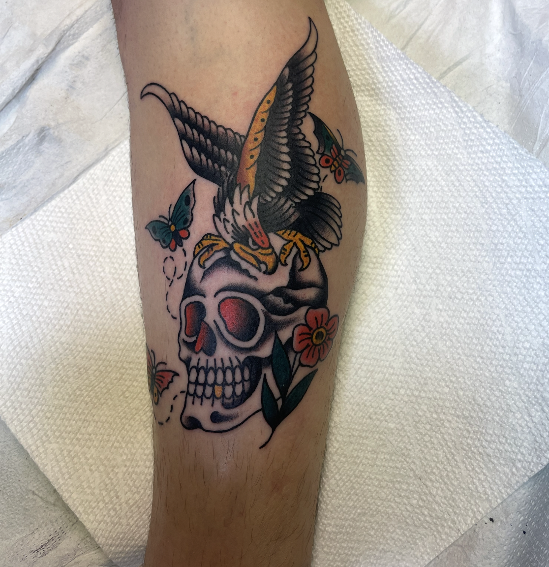Tattoo of a skull, a flying hawk, colorful butterflies, and flowers on a person's leg.