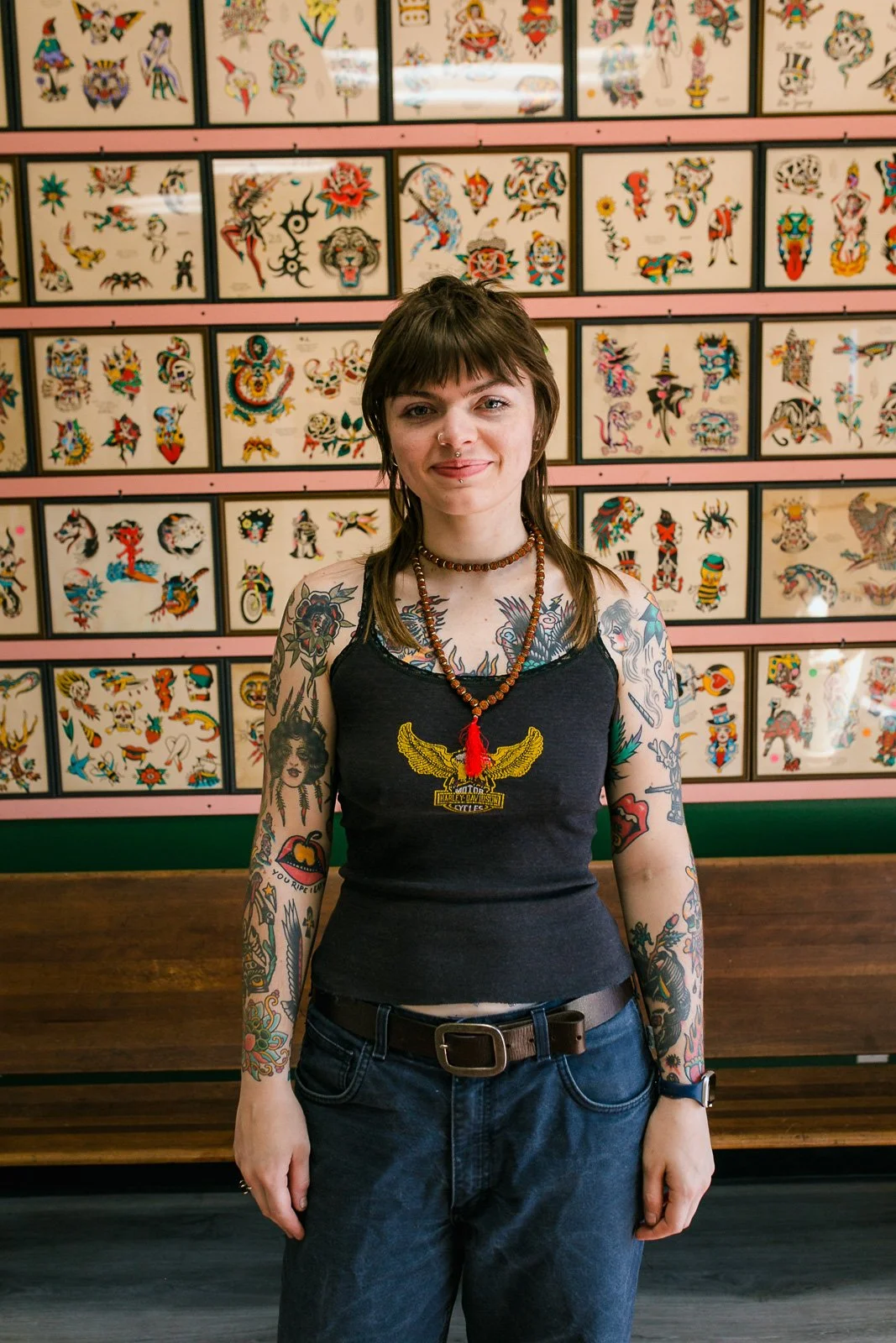 A woman with tattoos on her arms and chest, wearing a black tank top with a yellow eagle logo, standing in front of a wall decorated with colorful tattoo flash sheets.