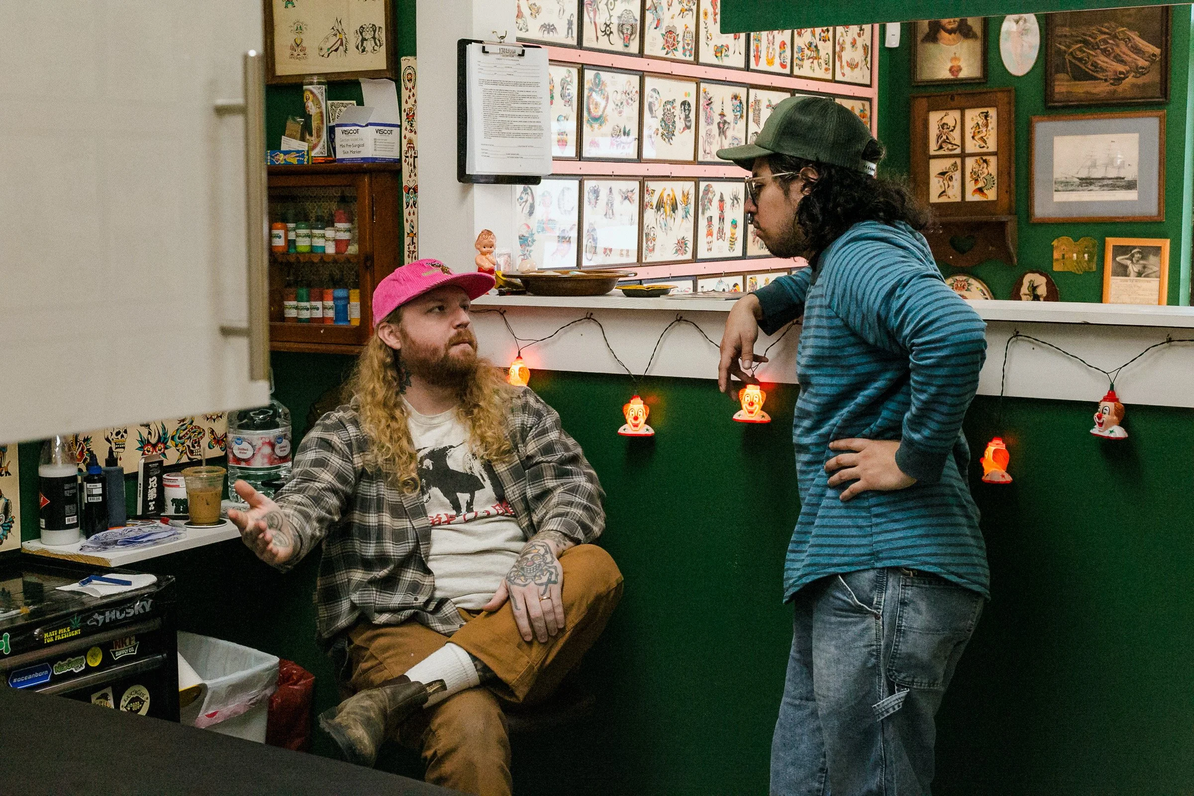 A man with blond hair, tattoos, and a beard is sitting at a table, wearing a pink hat and plaid shirt, talking to a woman standing in front of him. The woman has dark curly hair, glasses, and is wearing a green baseball cap and a blue striped shirt. 