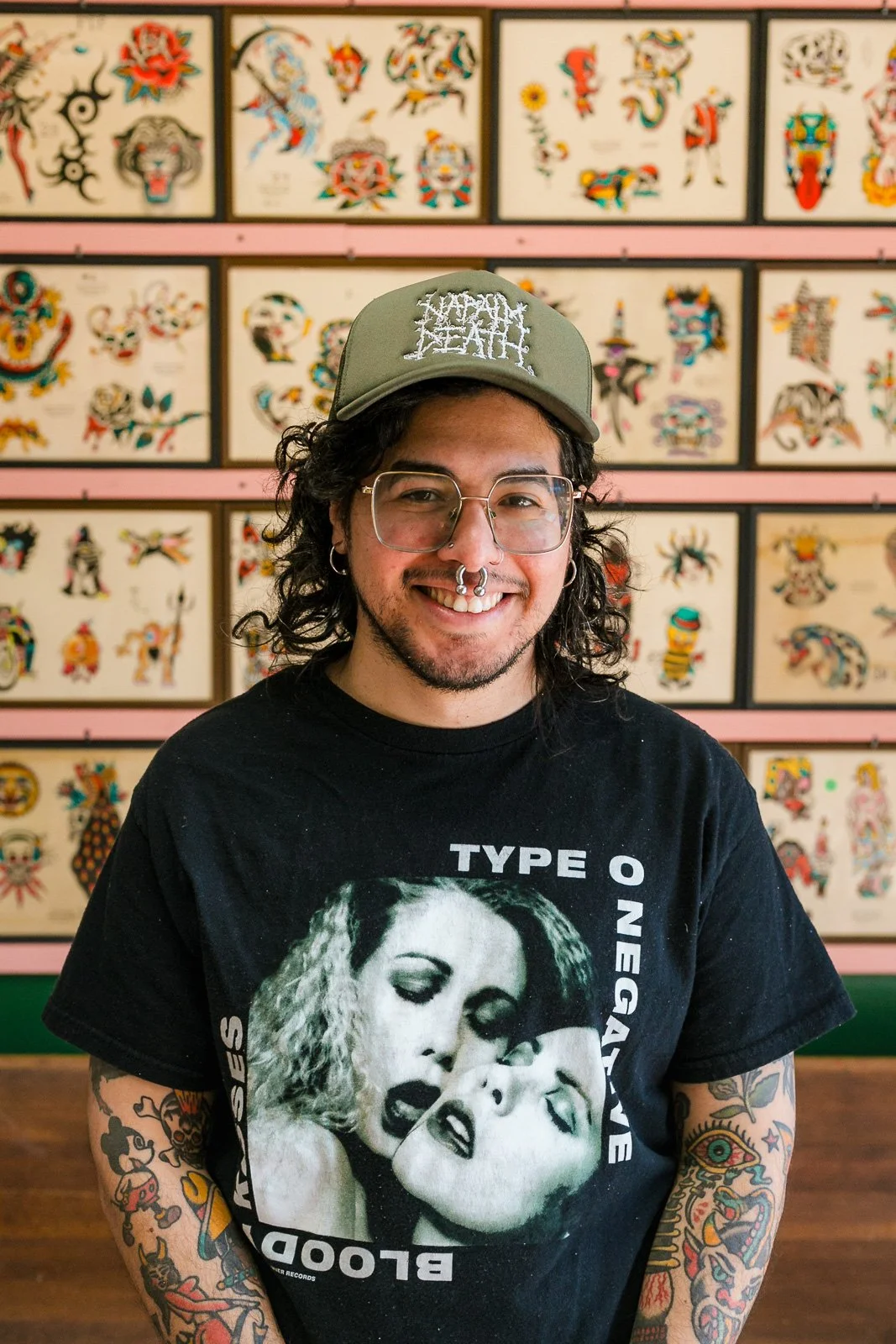 A man with curly black hair, glasses, and tattoos on his arms, wearing a black T-shirt with a Marilyn Monroe print and a green cap with the words 'HAPPY DEATH', smiling while standing in front of a wall decorated with colorful tattoo flash art.