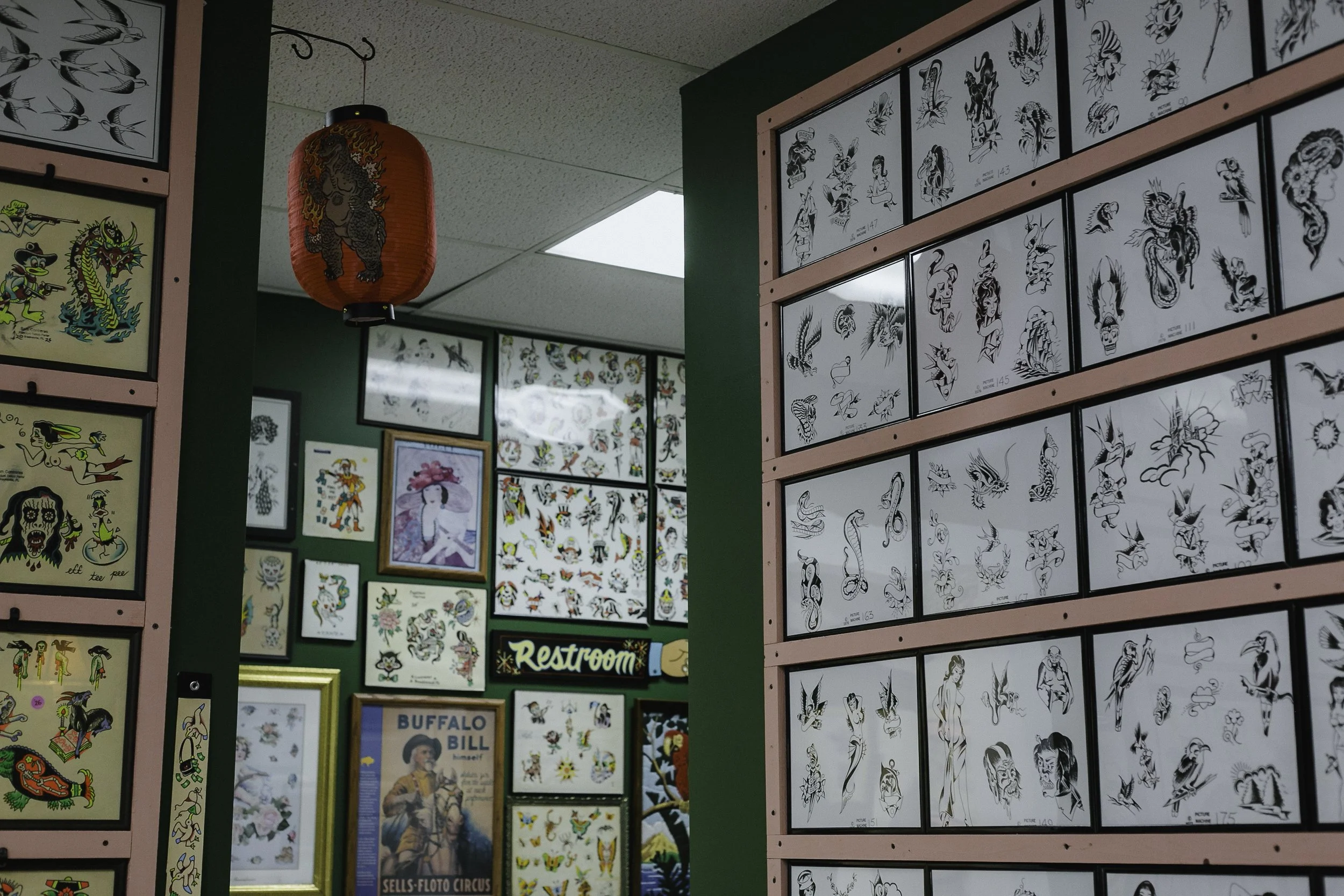 Wall decorated with various framed tattoo flash art, predominantly black and white, featuring traditional tattoo designs including animals, mythological figures, and pin-up style characters, along with a red Chinese lantern hanging from the ceiling.