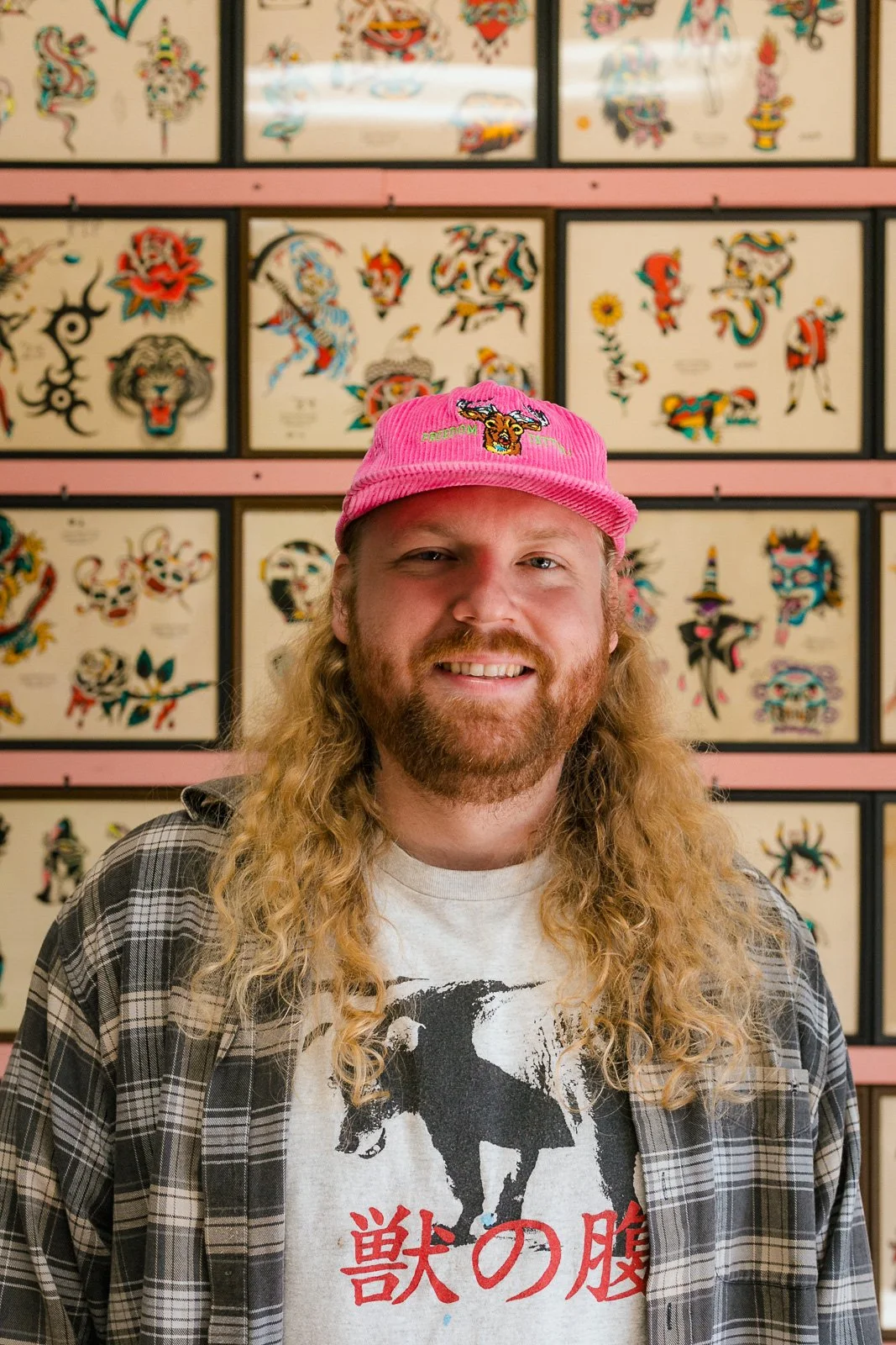 A man with long, curly blonde hair, a beard, and a smile, wearing a pink cap with tiger embroidery and a plaid shirt over a graphic T-shirt featuring a black dog and Japanese characters, standing in front of a wall decorated with colorful tattoo-style art of various characters and creatures.