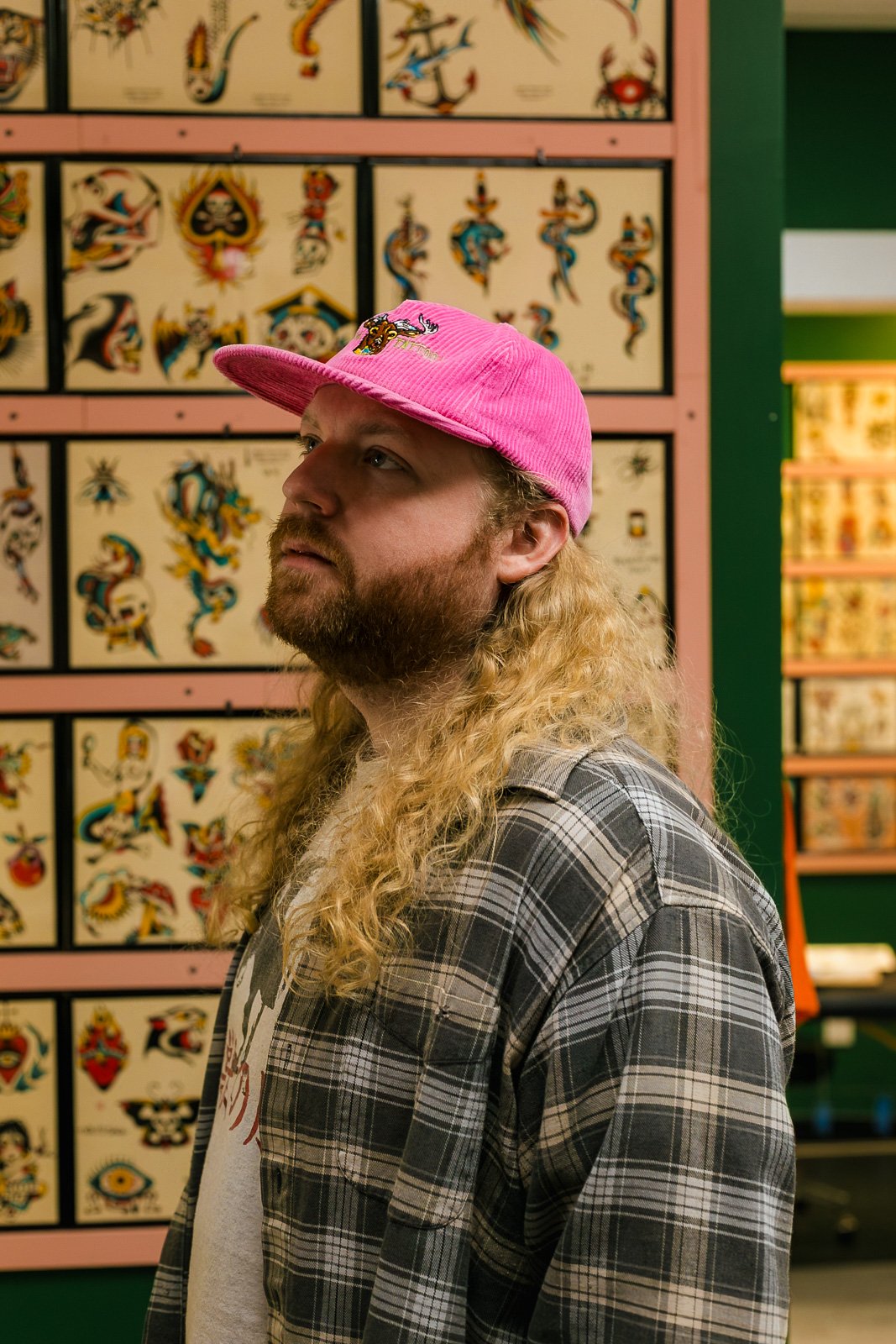 A man with long curly blond hair and a beard, wearing a pink cap and a gray plaid shirt, stands in front of a display of traditional tattoo flash art with various colorful tattoo designs.