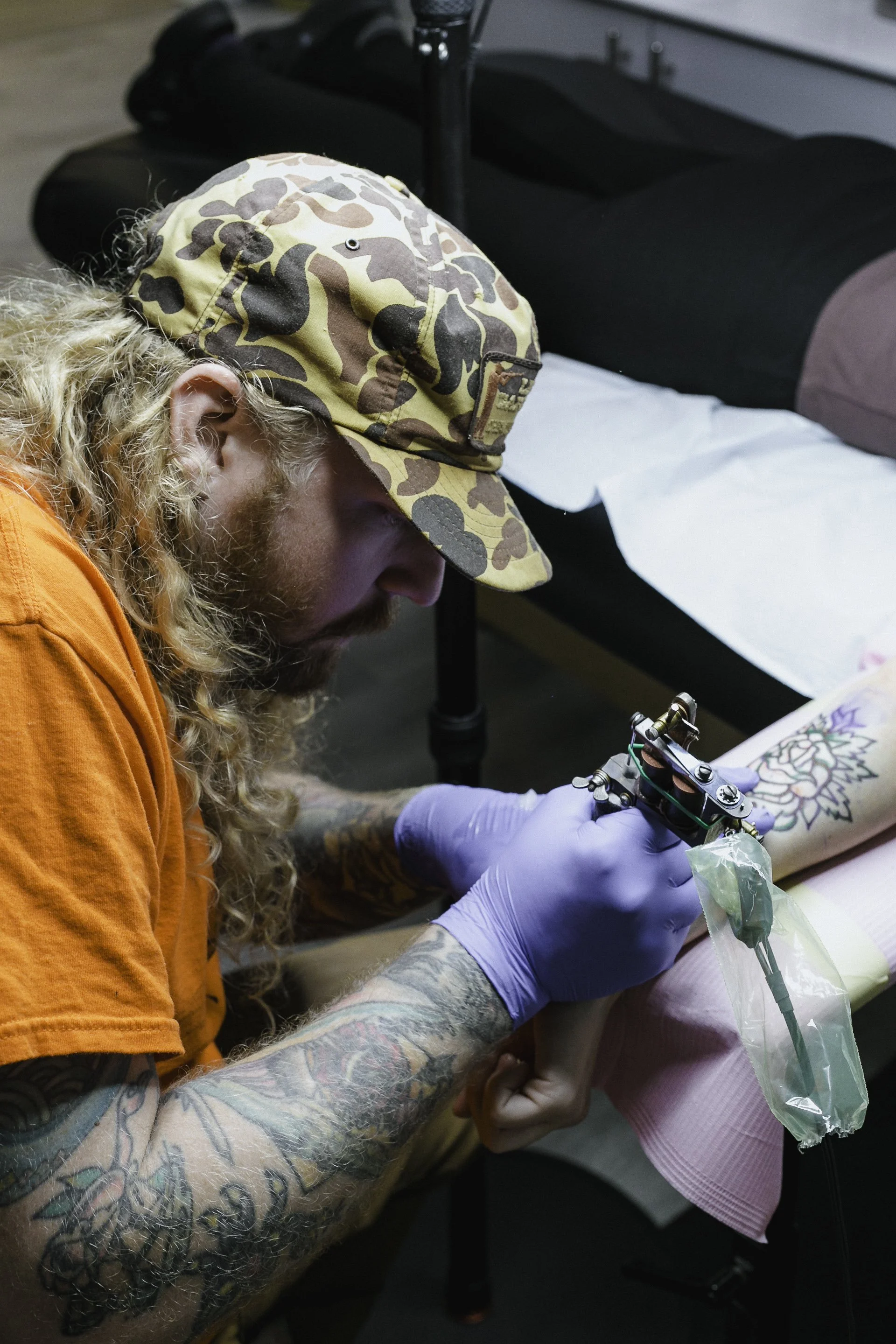 A man with tattoos on his arm and long curly hair, wearing an orange shirt and a camo hat, is tattooing a design on a person's arm while holding a tattoo machine covered with a plastic wrap. The person receiving the tattoo is wearing a pink sleeve.