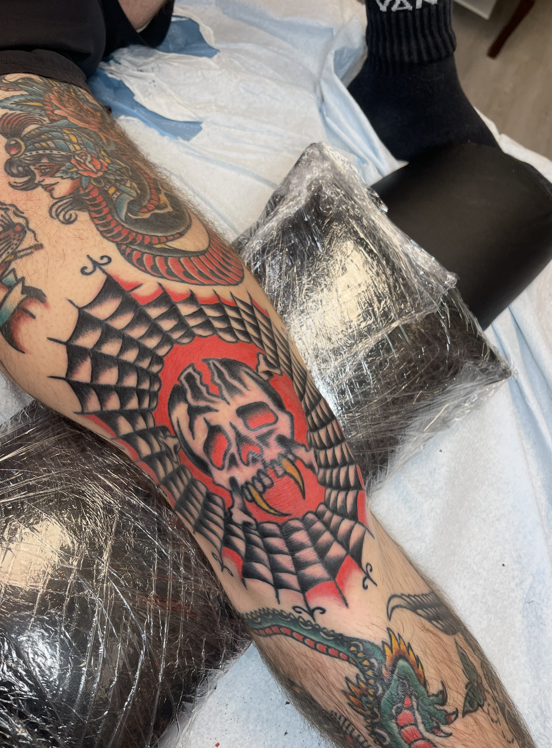 A tattoo artist's arm with colorful tattoo artwork, including a skull inside a spider web design, lying on a white sheet with tattoo equipment covered in plastic nearby.