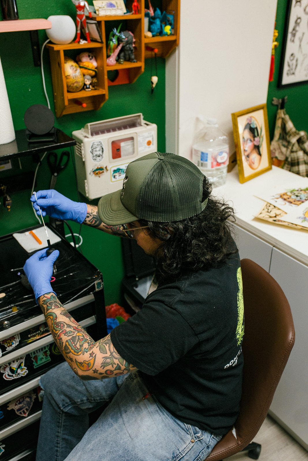 A person with curly hair, tattoos on their arms, wearing a black T-shirt, a green cap, and blue gloves, working with tools in a workshop or studio with shelves of toys and art on the walls.