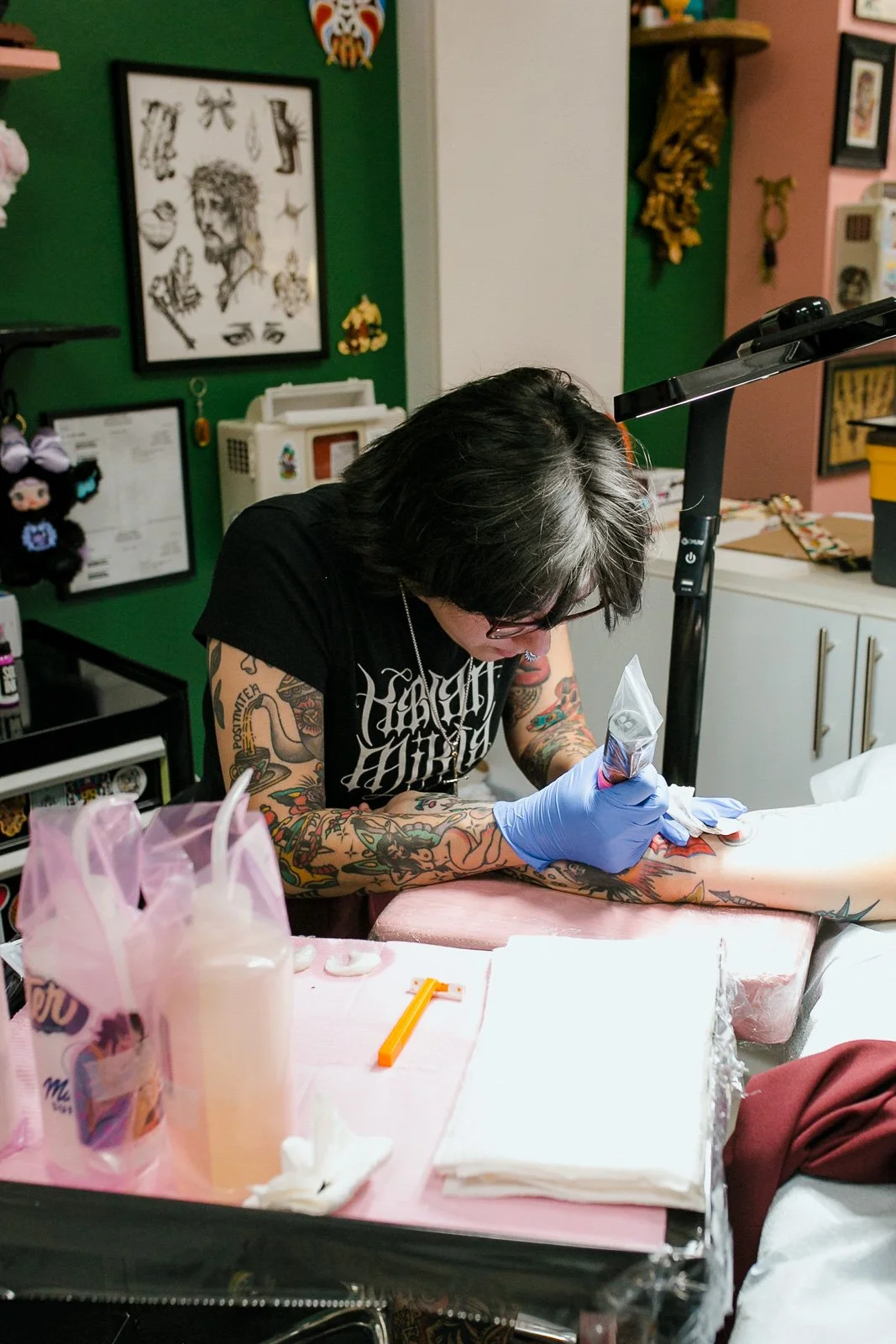 A tattoo artist wearing gloves and work glasses creating a tattoo on a person's arm in a tattoo studio with colorful artwork and decorations on the walls.