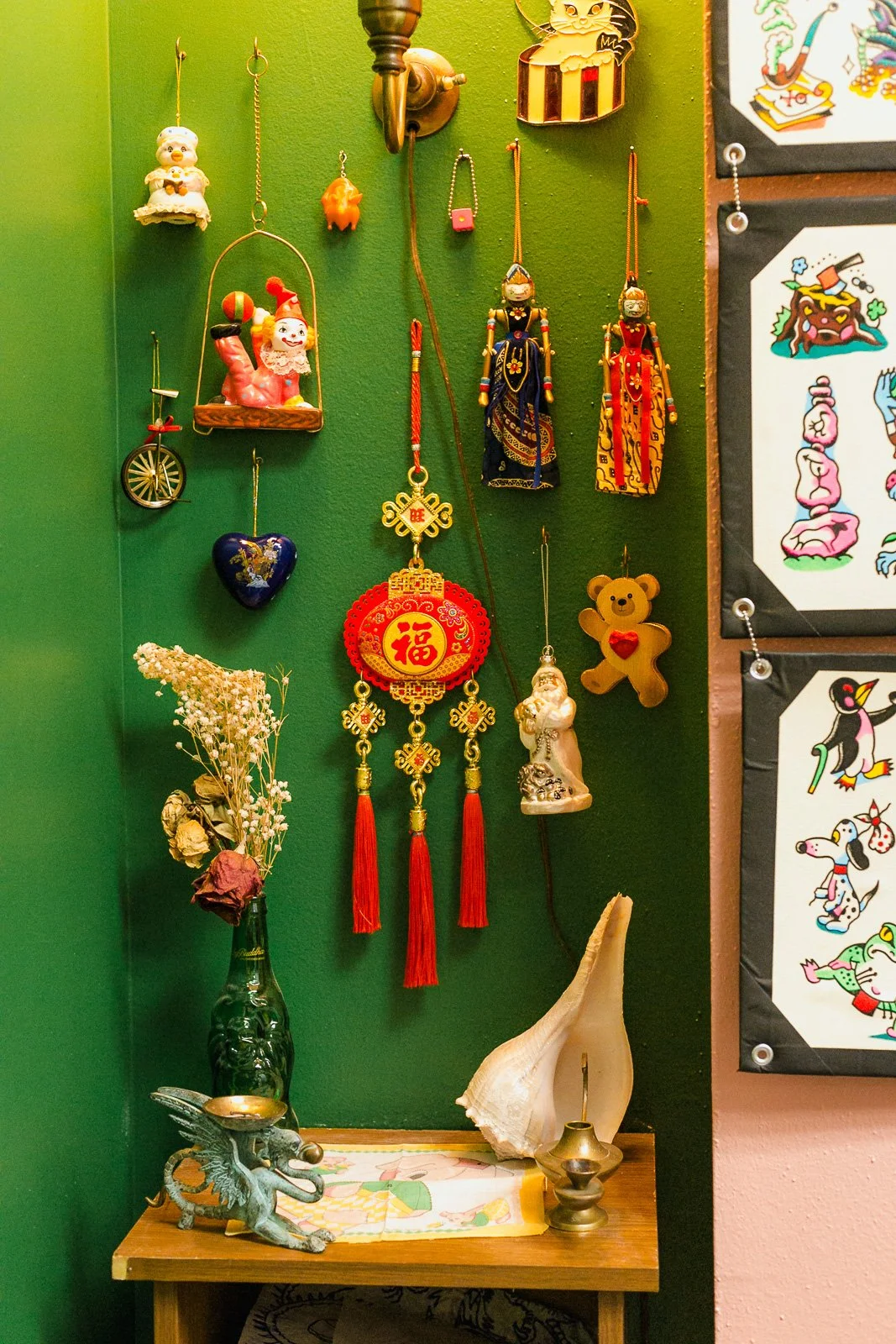Decorative Asian-themed ornaments hanging on a green wall, including figurines, lanterns, and jewelry, with a small wooden table below holding a vase with dried flowers, shells, and small sculptures.