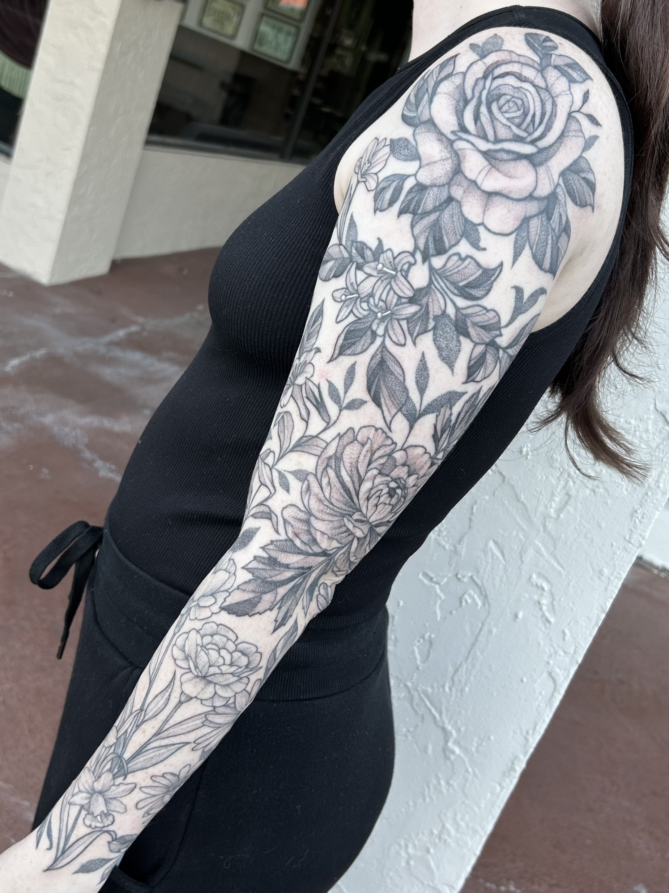 A person's arm with a large black and gray floral tattoo, including a rose and various other flowers, visible from the shoulder down to the wrist, wearing a sleeveless black top.