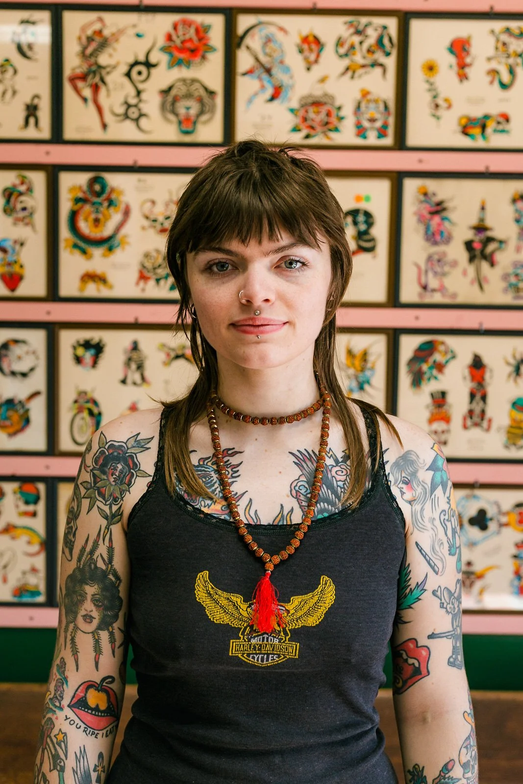 A young woman with short brown hair and tattoos on her arms and chest, wearing a black Harley Davidson tank top and beaded necklaces, stands in front of a wall covered with colorful tattoo flash art.