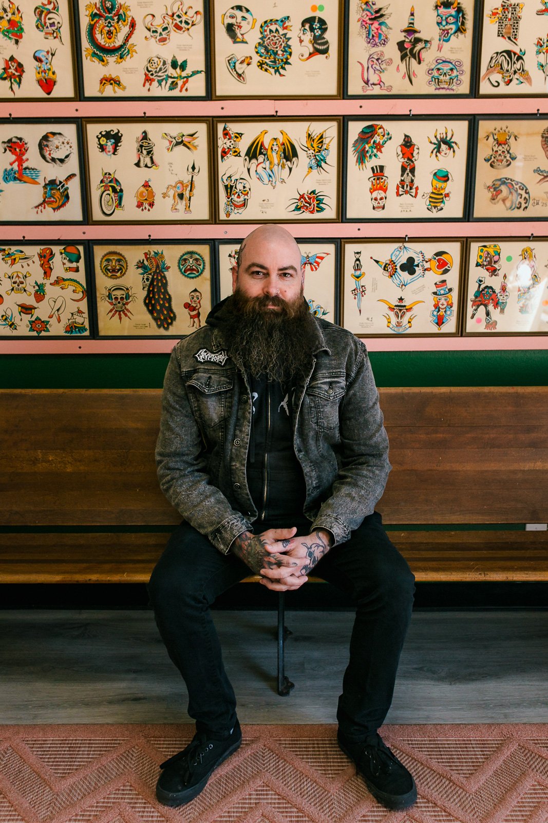 A man with a beard and tattoos sitting on a wooden bench in front of a wall filled with colorful tattoo flash art.