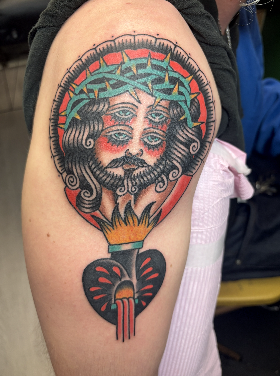 Tattoo of a face with multiple eyes and a crown of thorns, with a heart and flame below.