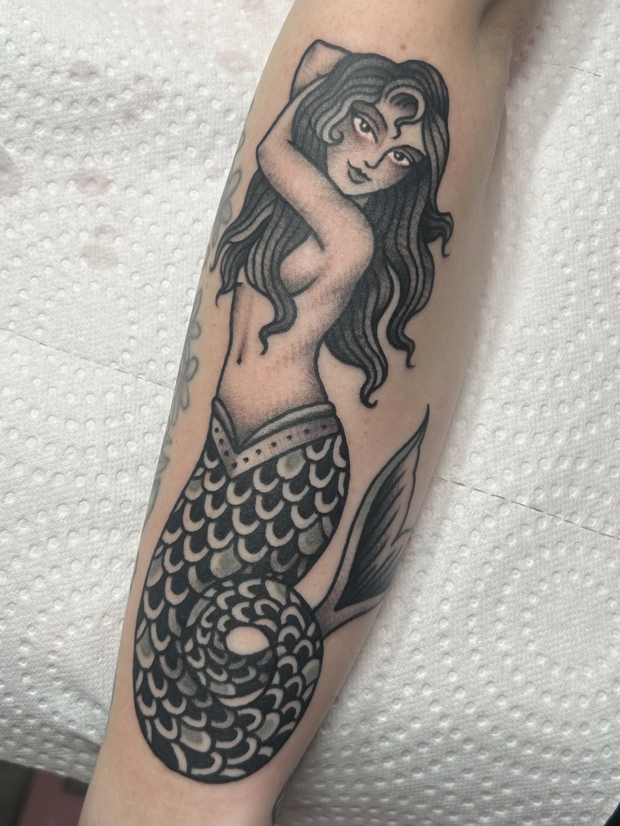 Black and gray tattoo of a mermaid on a person's arm, featuring detailed scales and flowing hair.