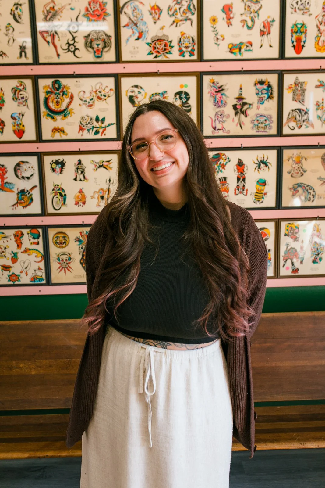 A woman with long wavy dark hair, glasses, and a black crop top, smiling in front of a colorful wall of traditional tattoo flash art.