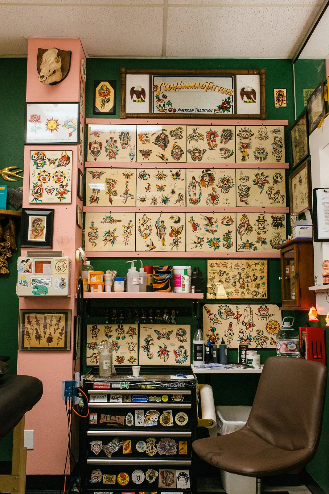 A wall decorated with numerous traditional tattoo flash sheets featuring colorful skulls, animals, and floral designs, with various tattoo supplies and a chair in the foreground.