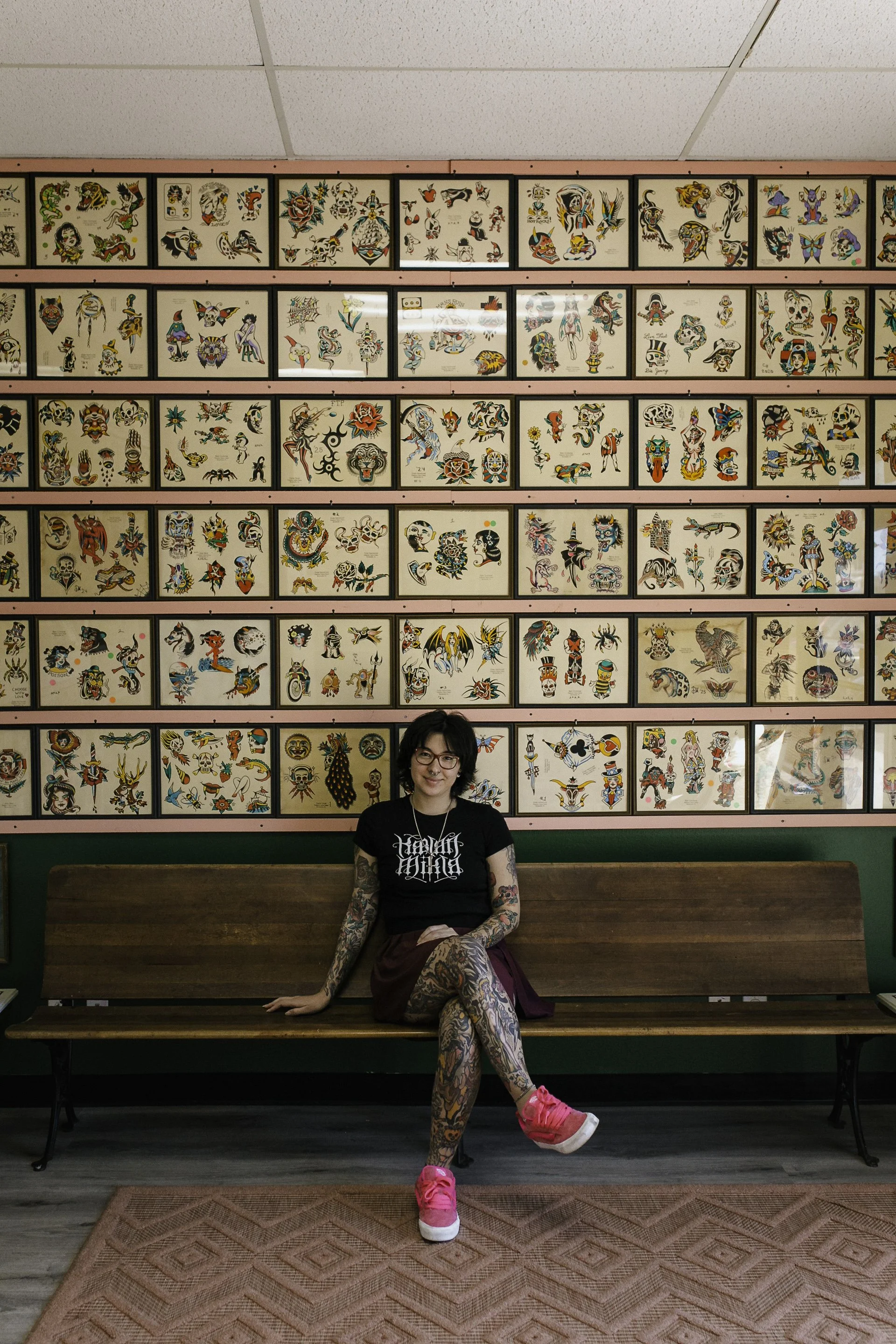 A woman with tattoos on her arms and legs sitting on a wooden bench in front of a wall filled with framed tattoo flash art. She is wearing a black t-shirt, patterned tights, bright pink sneakers, and has glasses and short dark hair.