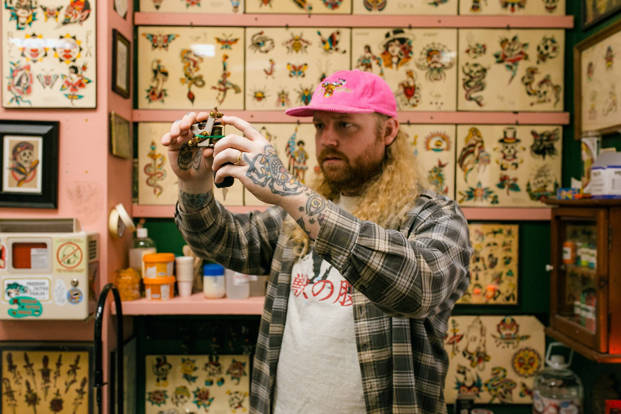 A man with long curly hair and tattoos on his arms wearing a pink cap and plaid shirt is examining a tattoo machine in a tattoo parlor decorated with tattoo artwork on the wall.
