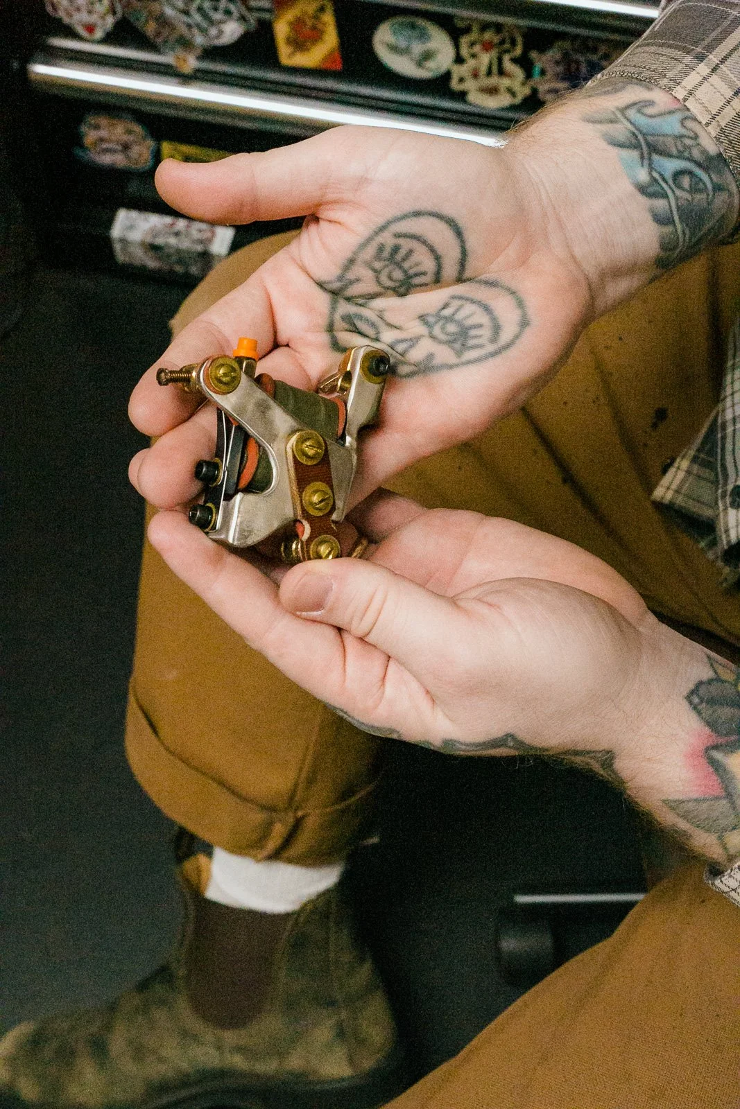 Person holding a tattoo machine with tattoos on their hand and wrist, in a tattoo studio.