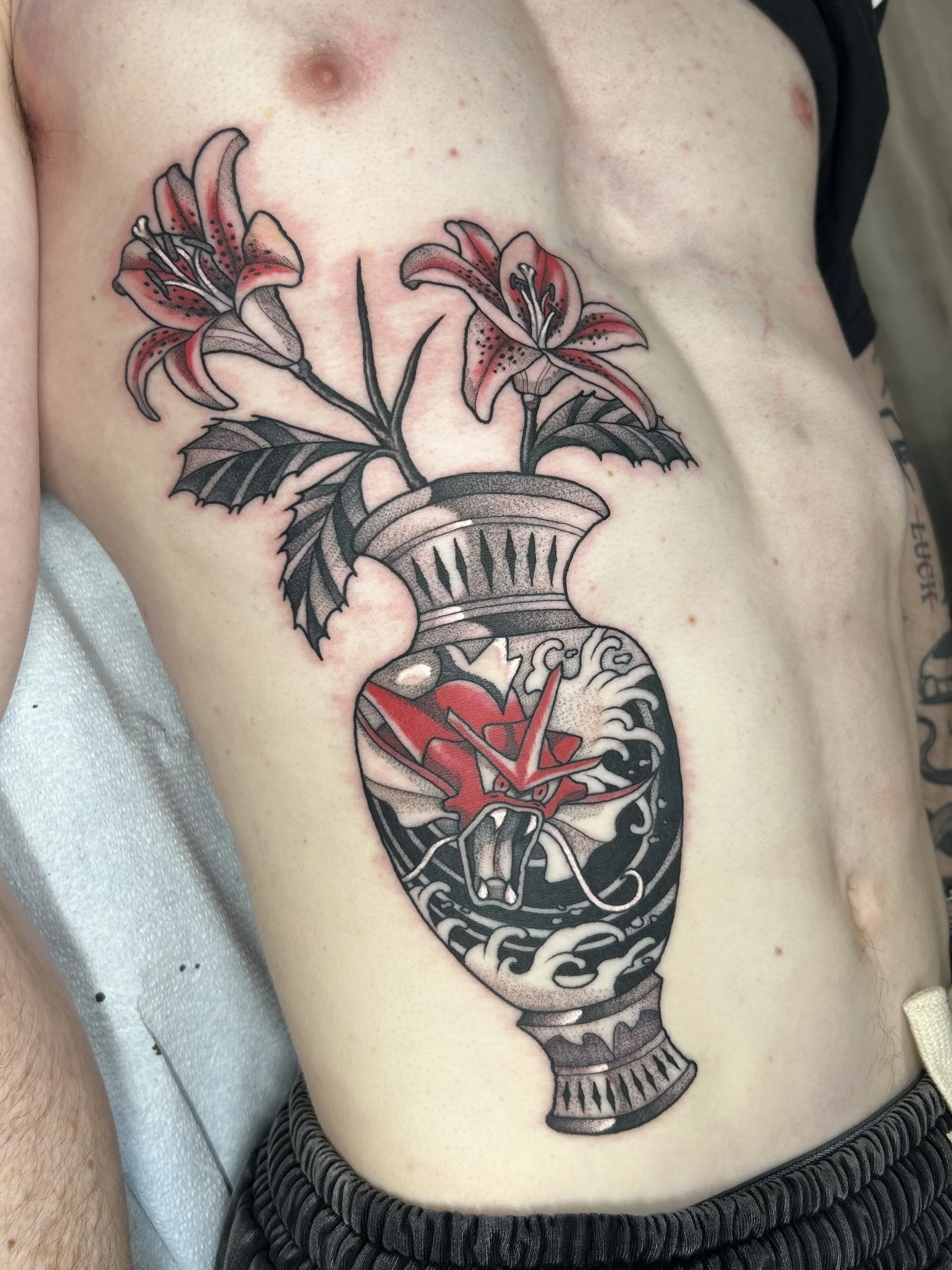 A tattoo on a person's side featuring a detailed lily flower in red and black ink, with leaves and a decorative vase. There's part of a tattoo that appears to be a clock or mechanical design below the lilies.