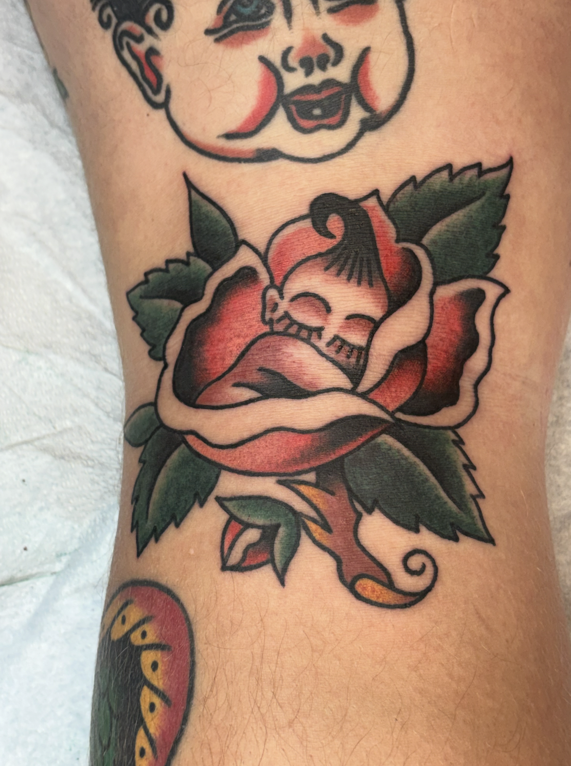 Tattoo of a smiling girl with a mohawk, surrounded by a red and green rose, depicted in a traditional tattoo style.