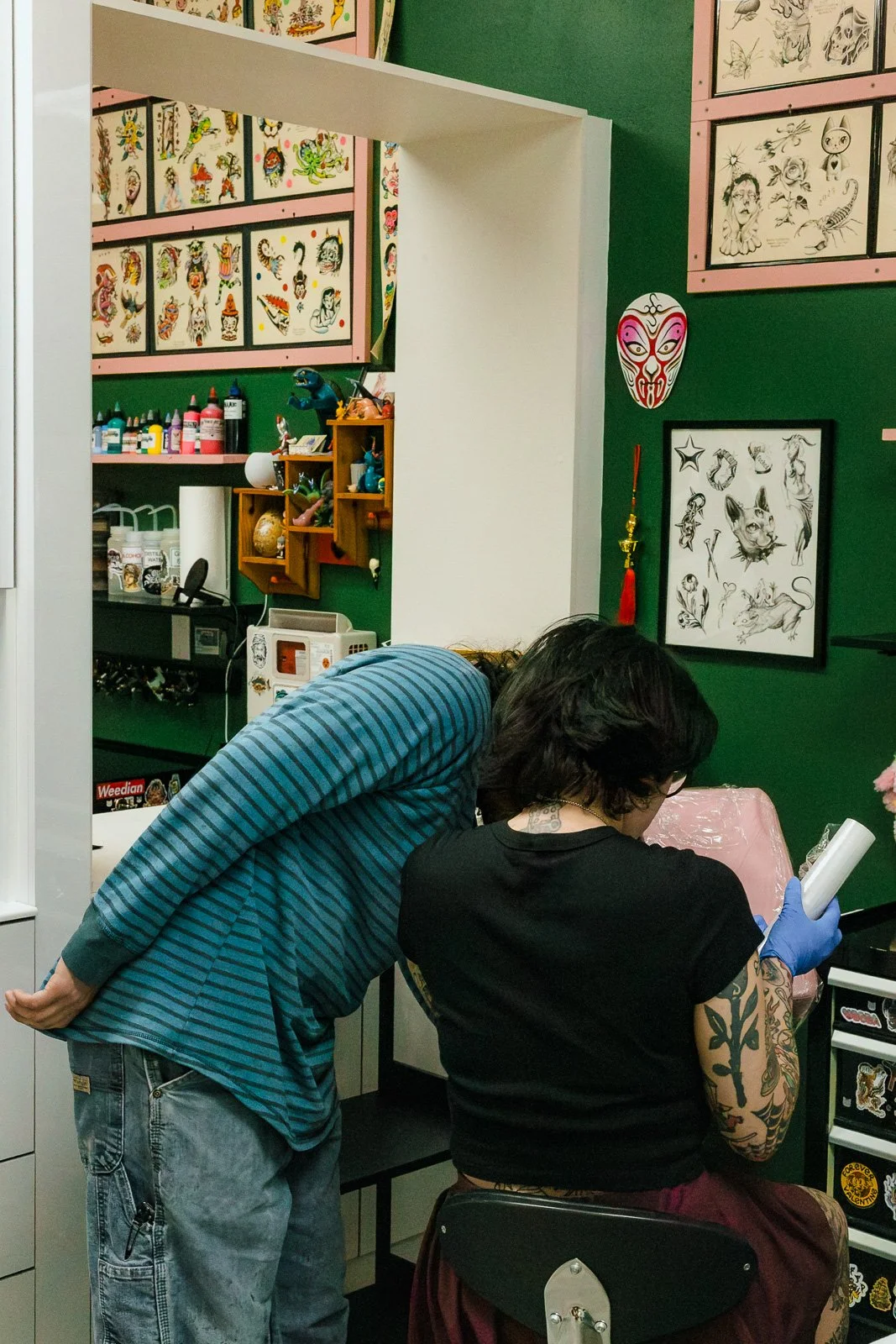 A tattoo artist with dark hair, glasses, and tattoos on her arms working on a client at a tattoo shop. The client has short dark hair and glasses, and is wearing a black shirt. The shop has green walls with framed tattoo designs and various artwork, 