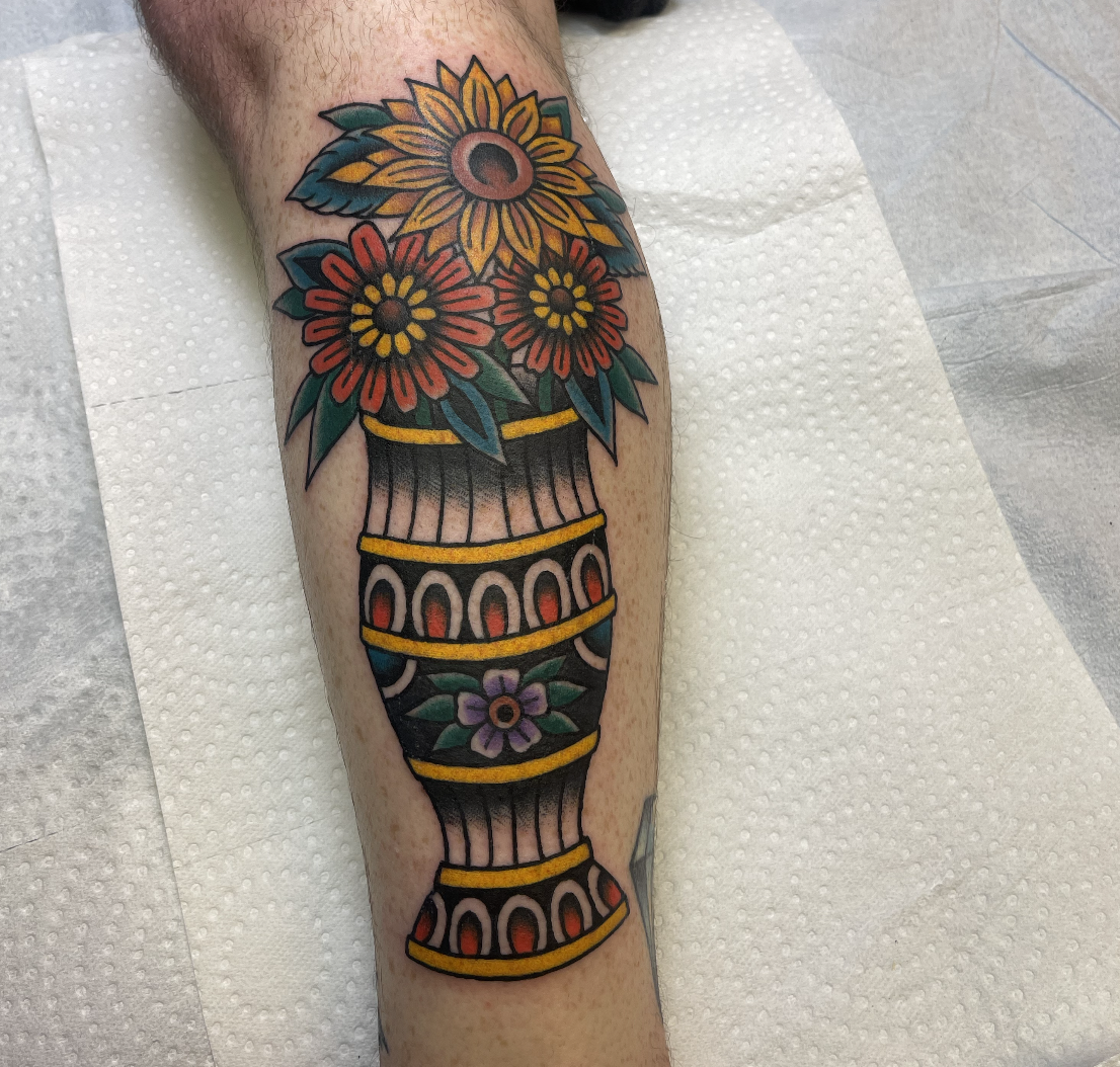 Colorful tattoo of a bouquet of sunflowers and daisies in a decorative vase on a person's leg.