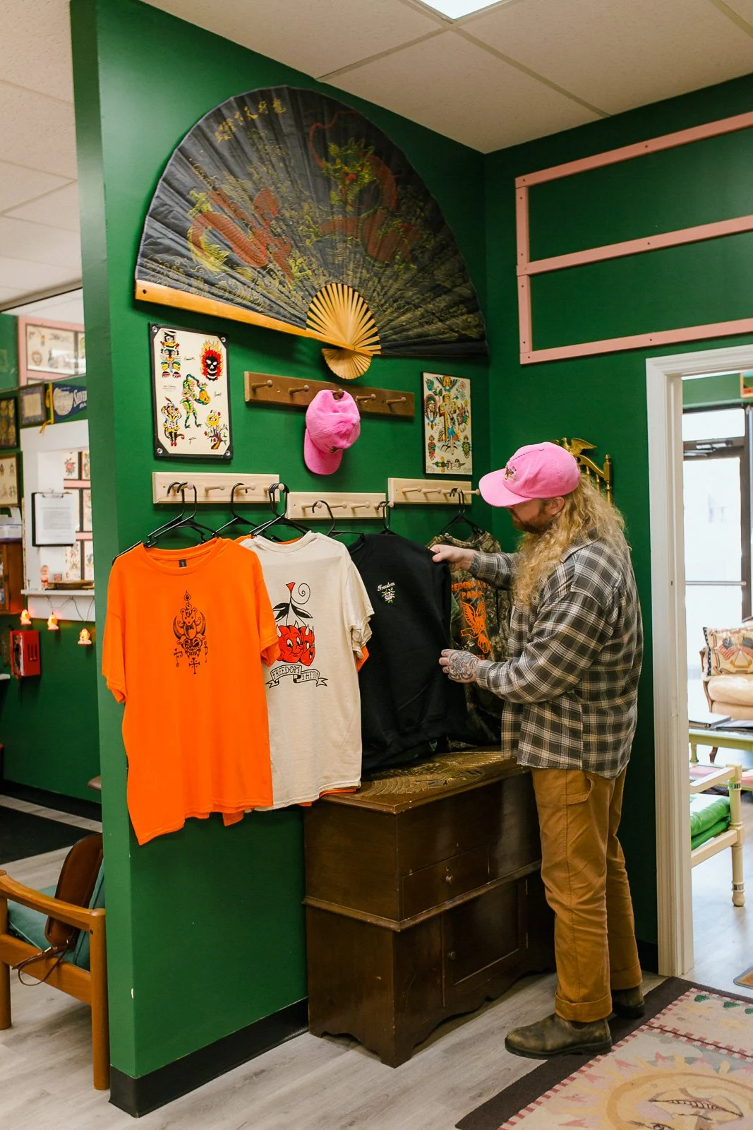 A man with long blonde hair, a plaid shirt, and a pink cap looks at black and orange graphic t-shirts on display in a store with green walls decorated with Asian art and a large black fan.