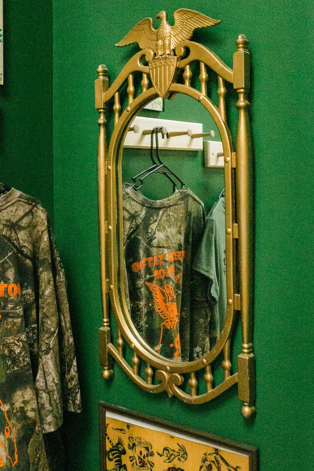 A gold-framed mirror with an eagle crest on top mounted on a green wall. Reflects a camouflage T-shirt with orange text and a tiger graphic hanging on a hanger.