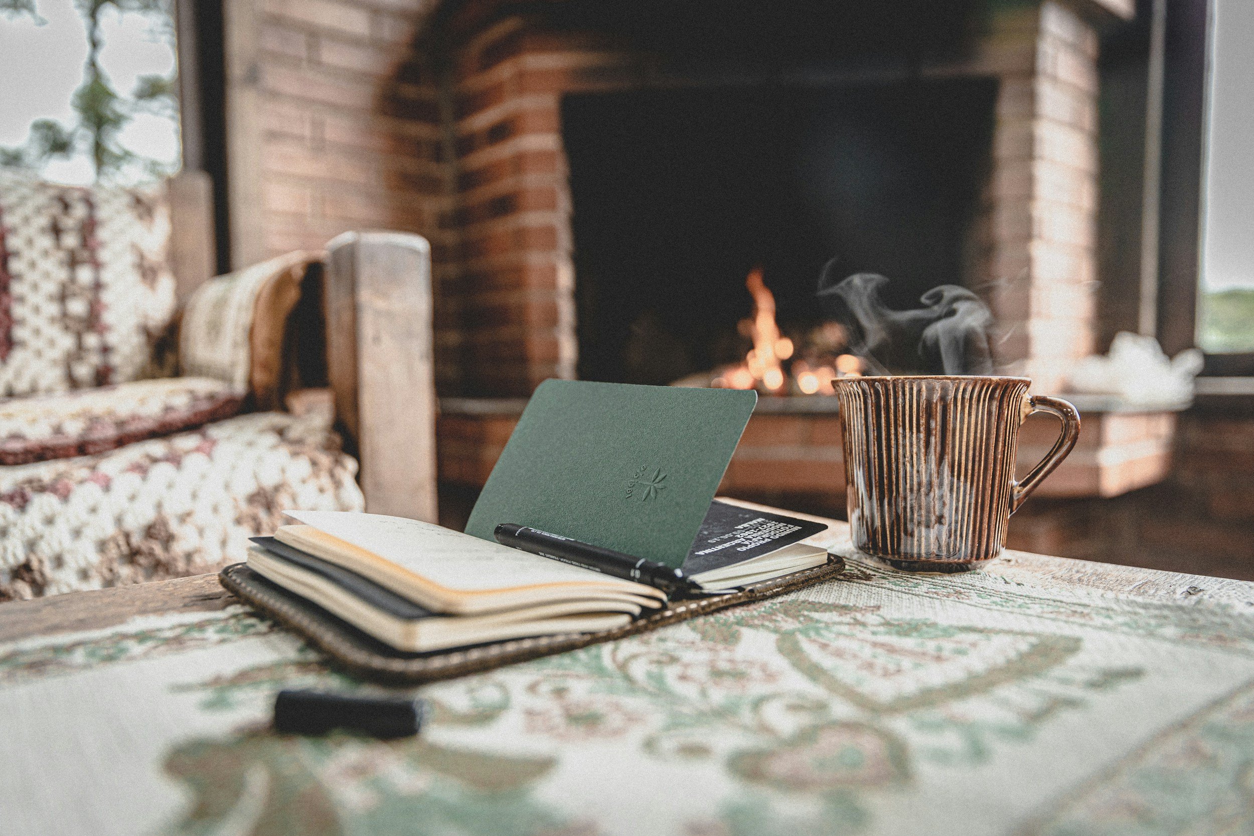 A cozy indoor scene with a steaming mug on a table, an open notebook with a pen, a closed book, and a green card or journal. Fireplace with a fire burning is in the background.