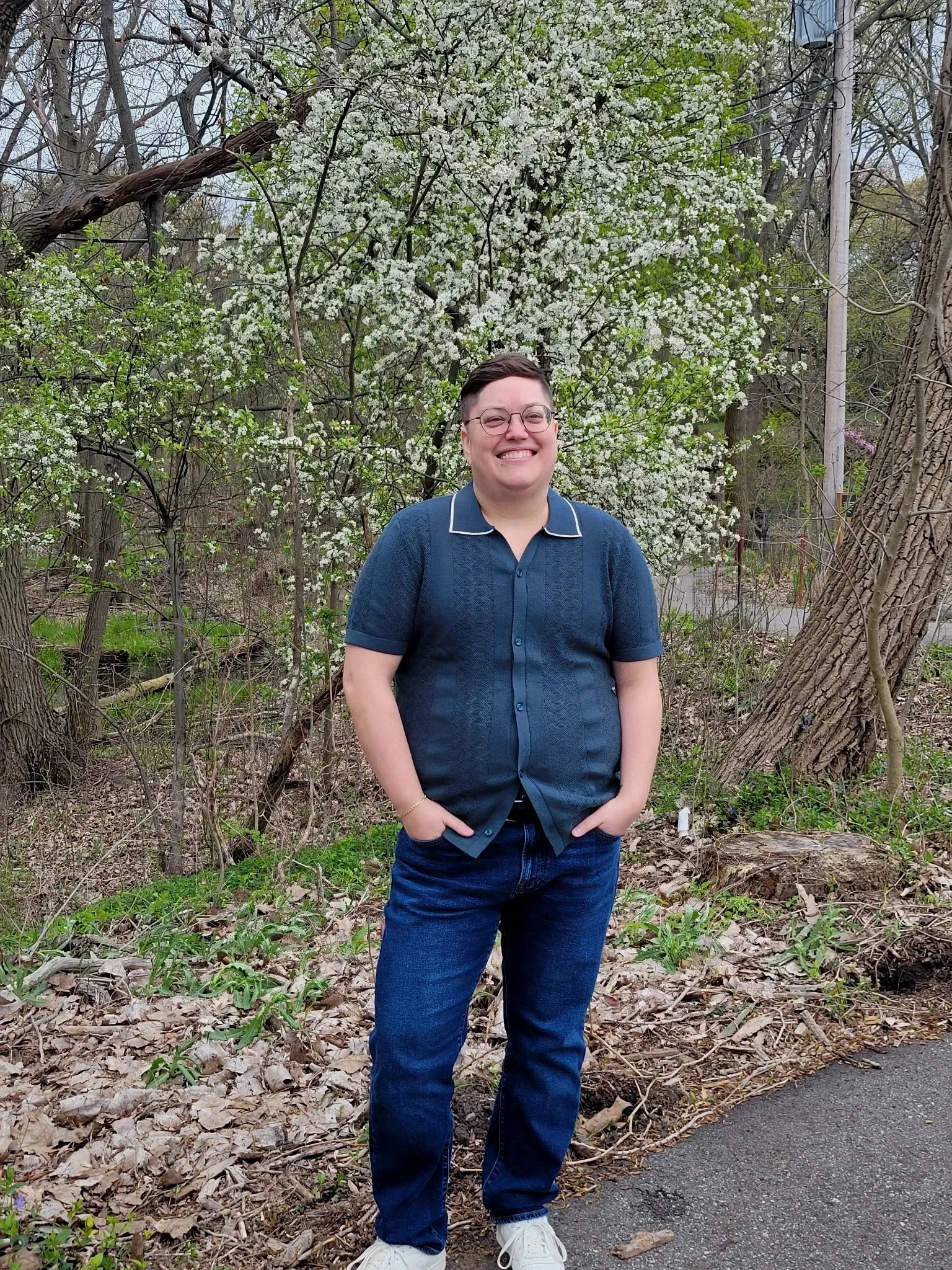 A person standing outdoors in front of a flowering tree, wearing a navy blue shirt, jeans, and white sneakers, smiling with hands in pockets.