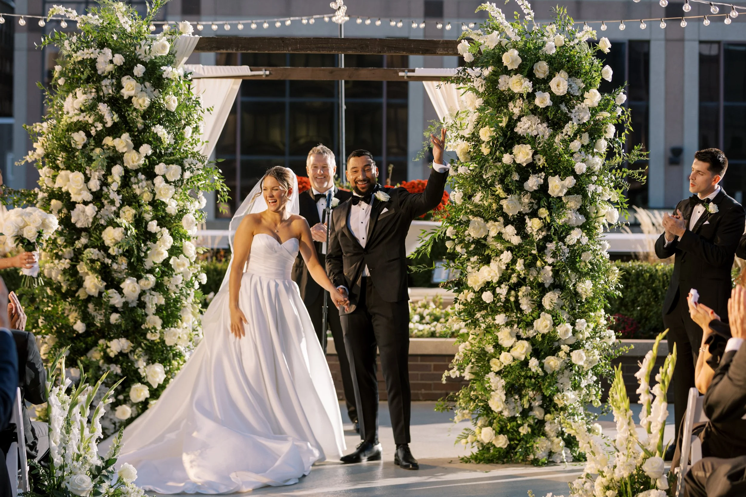 Outdoor rooftop wedding ceremony on the 5th-floor terrace at JPS Events at Regions Tower, featuring a lush white rose arch by our in-house florists, 360-degree downtown Indy skyline views, and built-in day-of coordination.