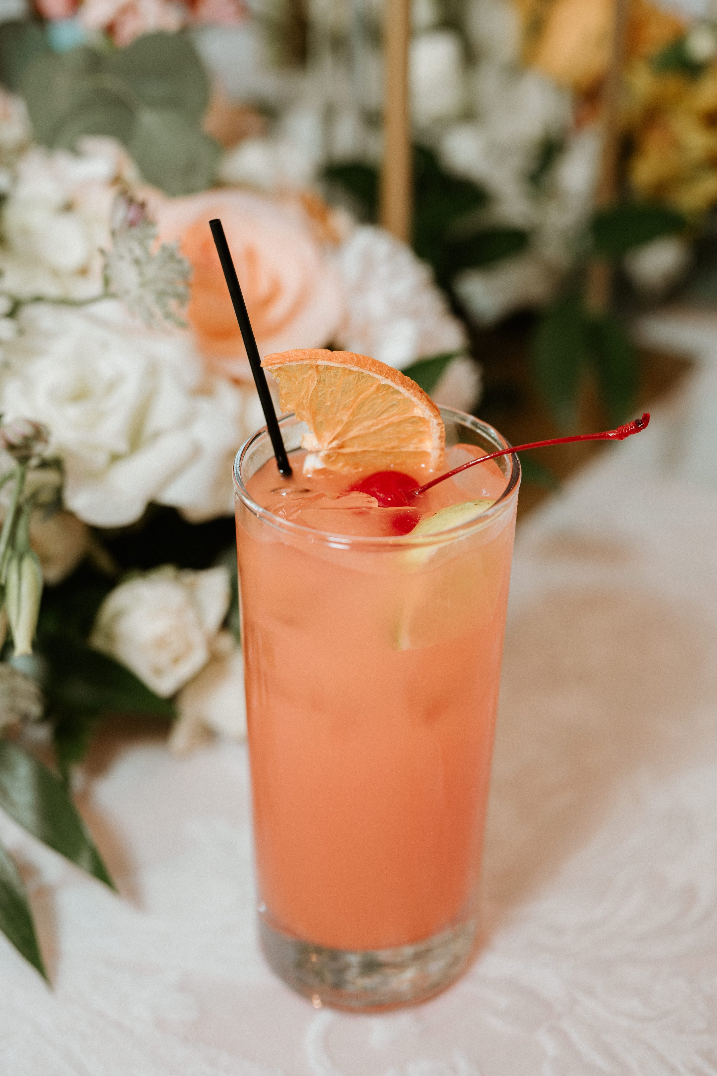 A pink cocktail drink garnished with a slice of orange and a cherry, with a black straw, on a table with a floral background.