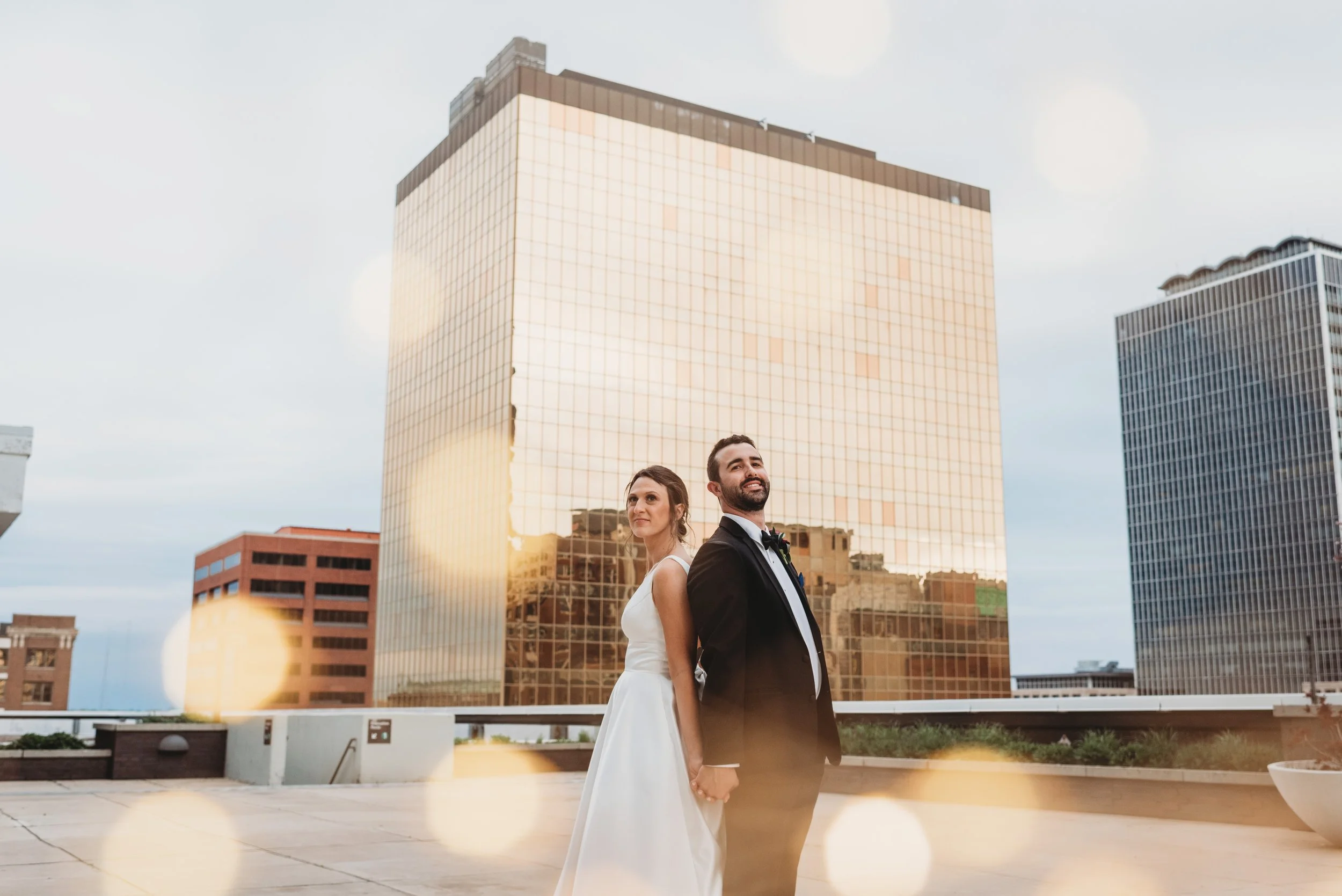 A newlywed couple in wedding attire standing outdoors holding hands in front of tall glass buildings, with sunlight reflecting off the buildings and soft lens flare.
