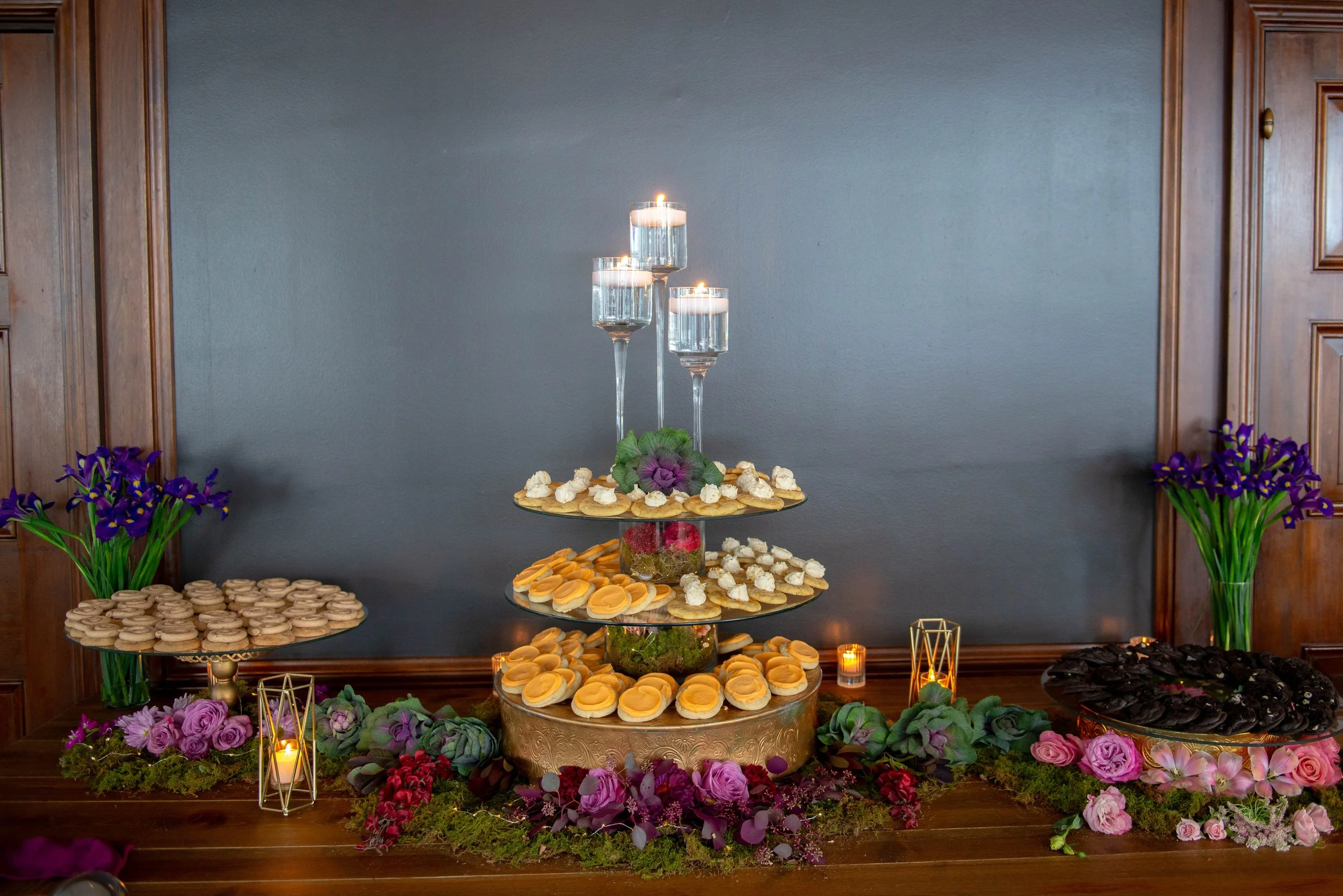 A dessert table with lit candles, flowers, and various treats on tiers, set against a blue wall and wooden panels.