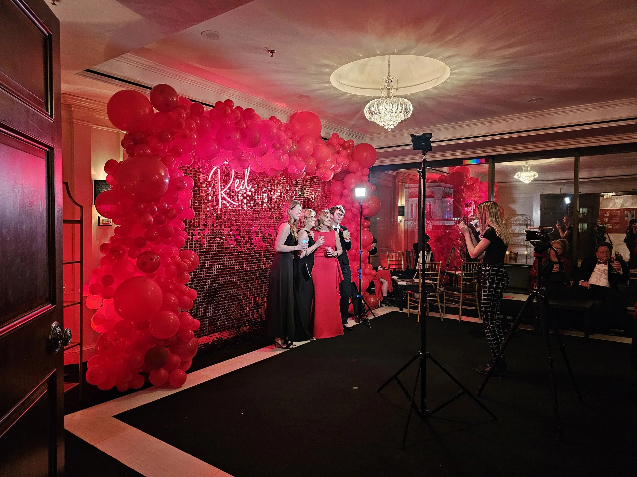 A group of four women dressed in formal attire standing in front of a red balloon wall with the word 'Red' illuminated in neon. The setting appears to be a party or celebration event in a decorated indoor space with chandeliers and guests.