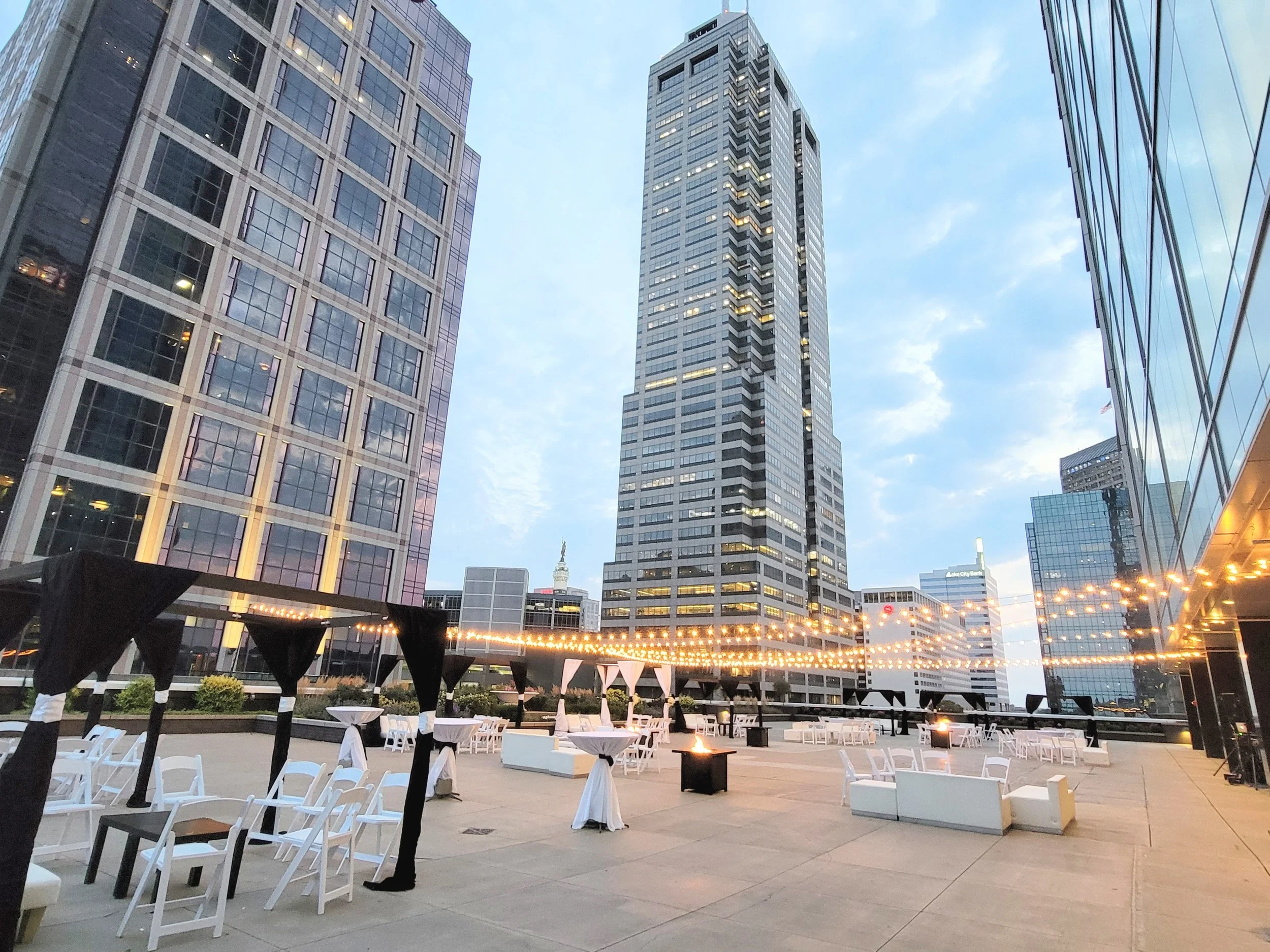 Outdoor rooftop area with white tables and chairs, string lights, and modern high-rise buildings in the background during early evening.